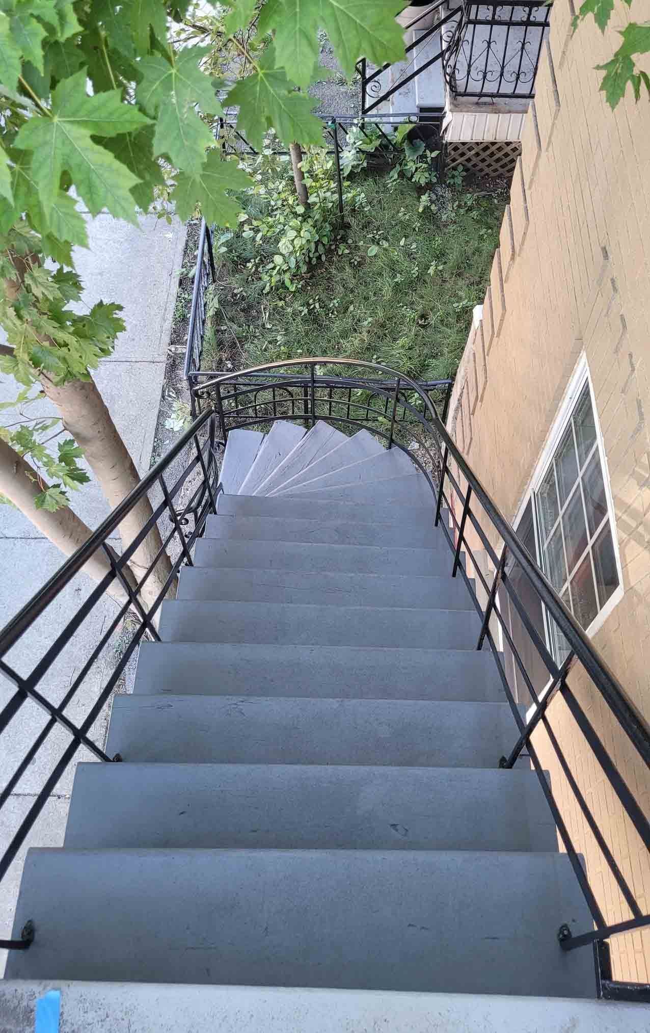 Steep outdoor staircase with black railings beside a brick building and greenery below