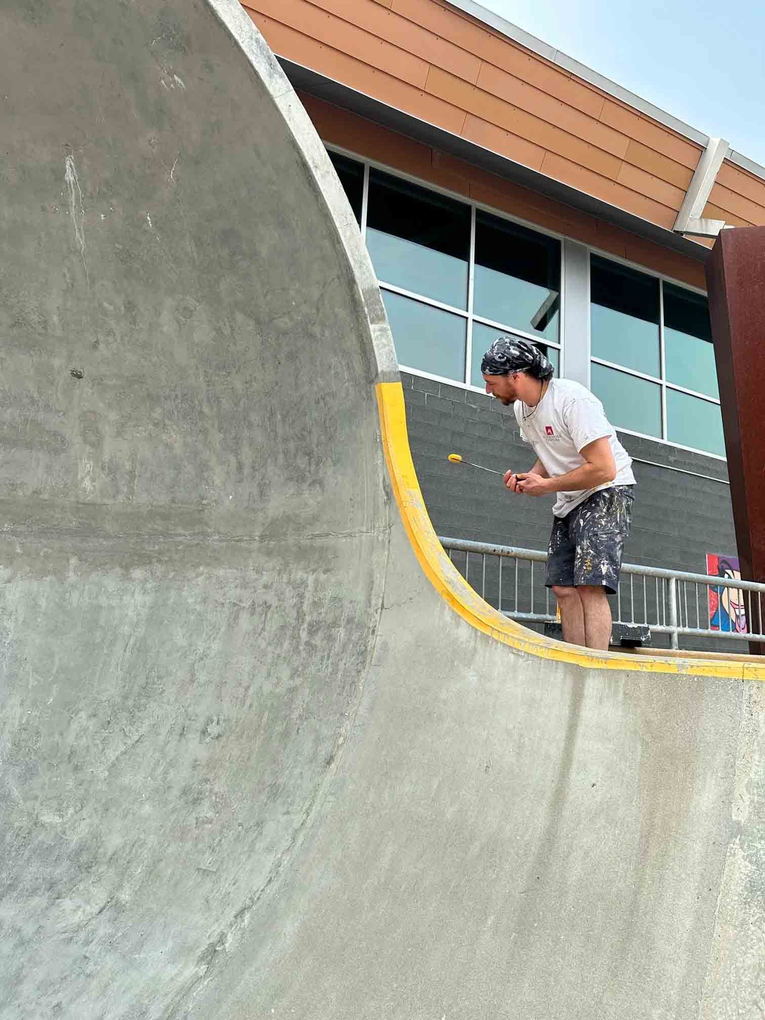 Man standing on a skateboard ramp beside a yellow edge, looking at a phone outdoors.