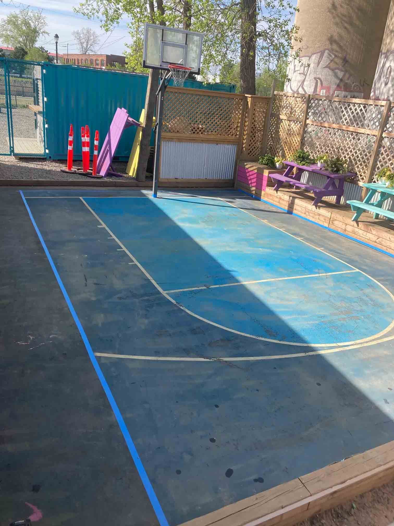 Outdoor basketball half court with blue lines, a hoop, and colorful bench seating beside a fenced yard.