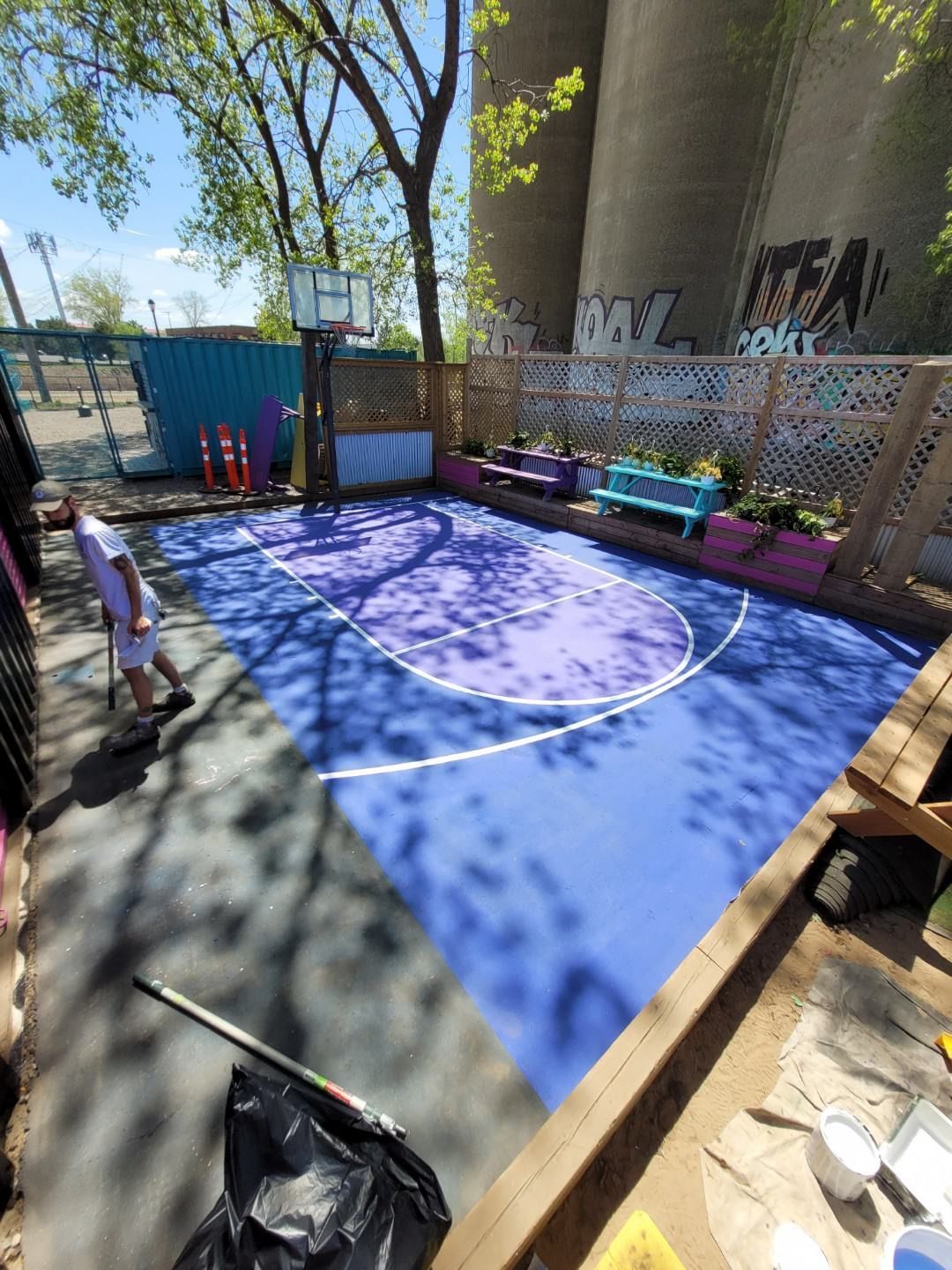 Blue outdoor sports court under a bridge, with a fenced play area and a child standing at left.