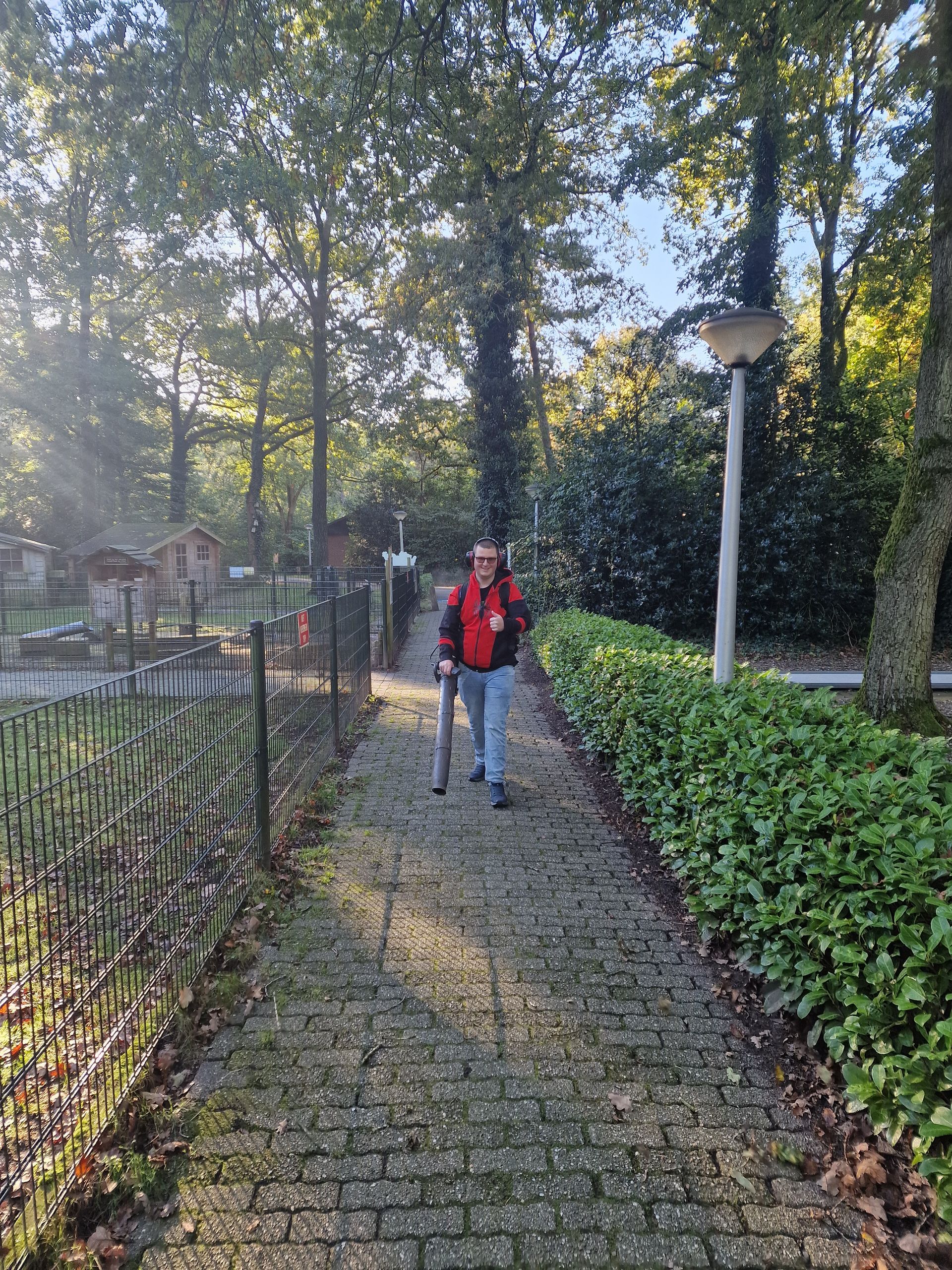 Een persoon loopt met een wandelstok over een stenen pad in een park. Bomen en struiken omringen het pad.