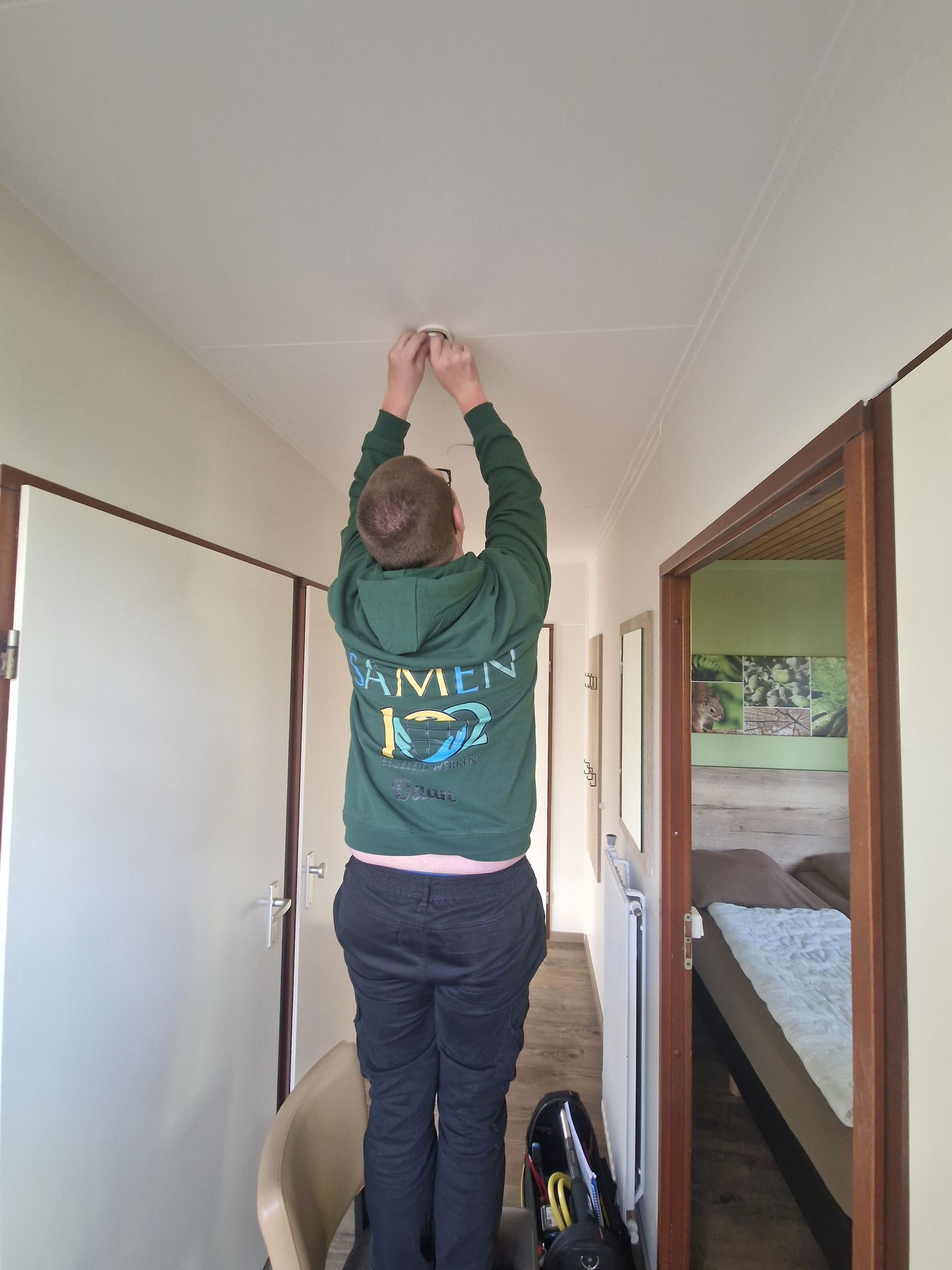 Persoon in groene hoodie, staand op een stoel, bezig met plafondwerkzaamheden in een gang.