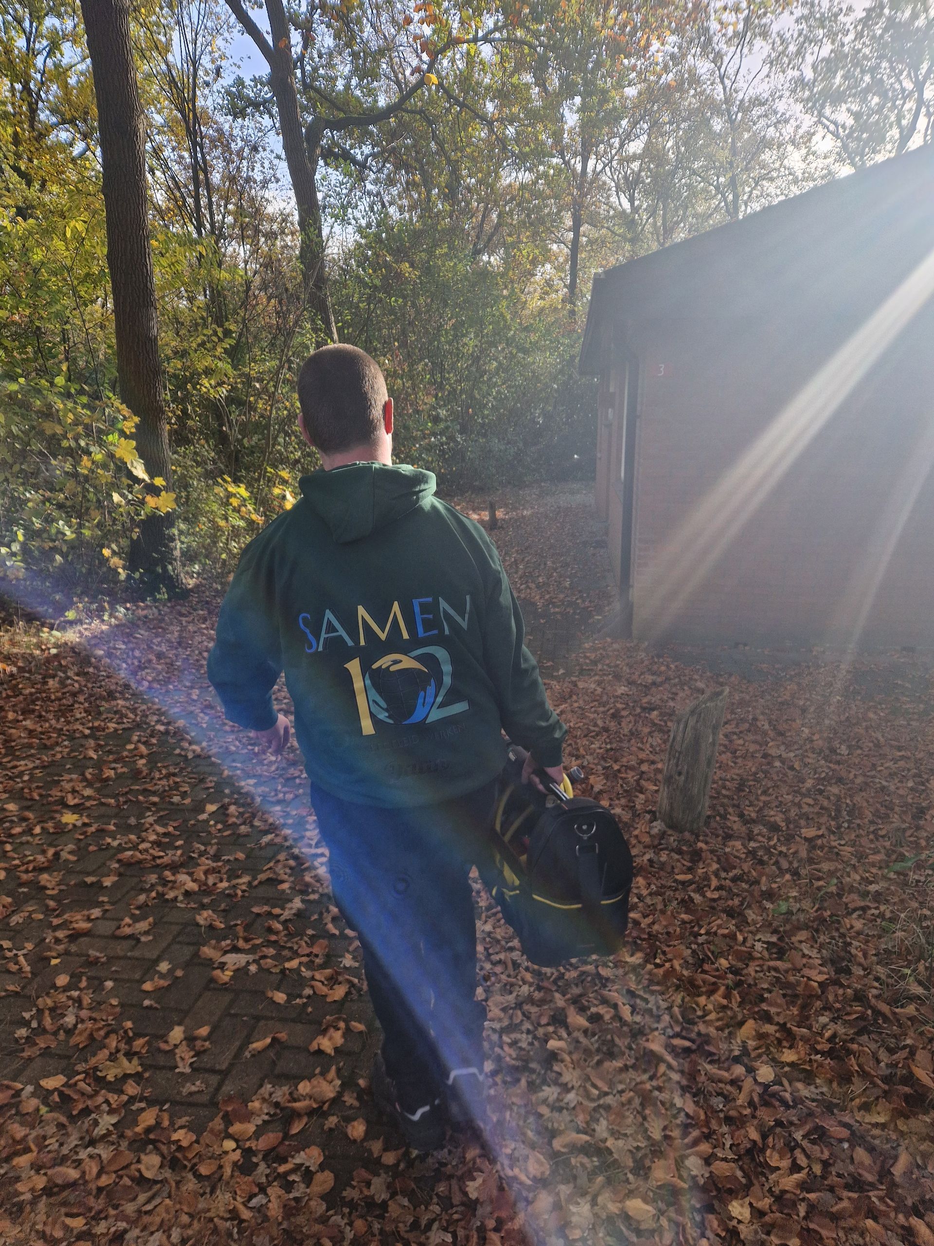 Een persoon in een groene hoodie loopt over een met bladeren bedekt pad en houdt een tas vast. 