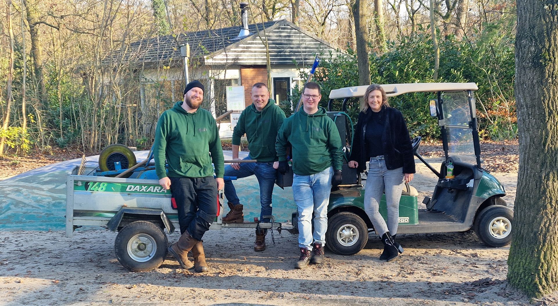 Vier personen in groene hoodies poseren bij een golfkarretje en een klein gebouw in een bosrijk gebied.