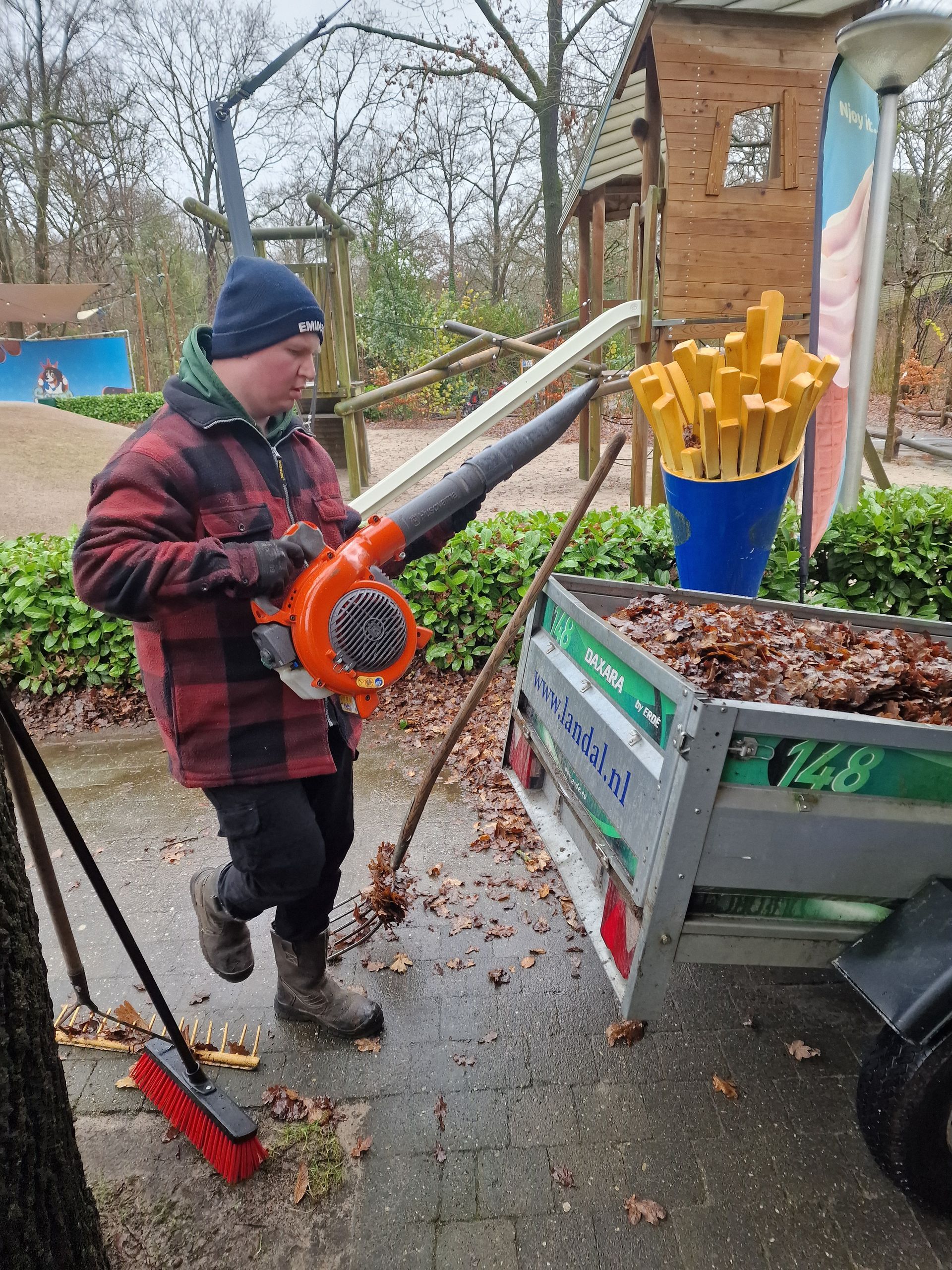 Een man gebruikt een bladblazer om bladeren in een aanhanger te vegen, vlakbij een speeltuin.
