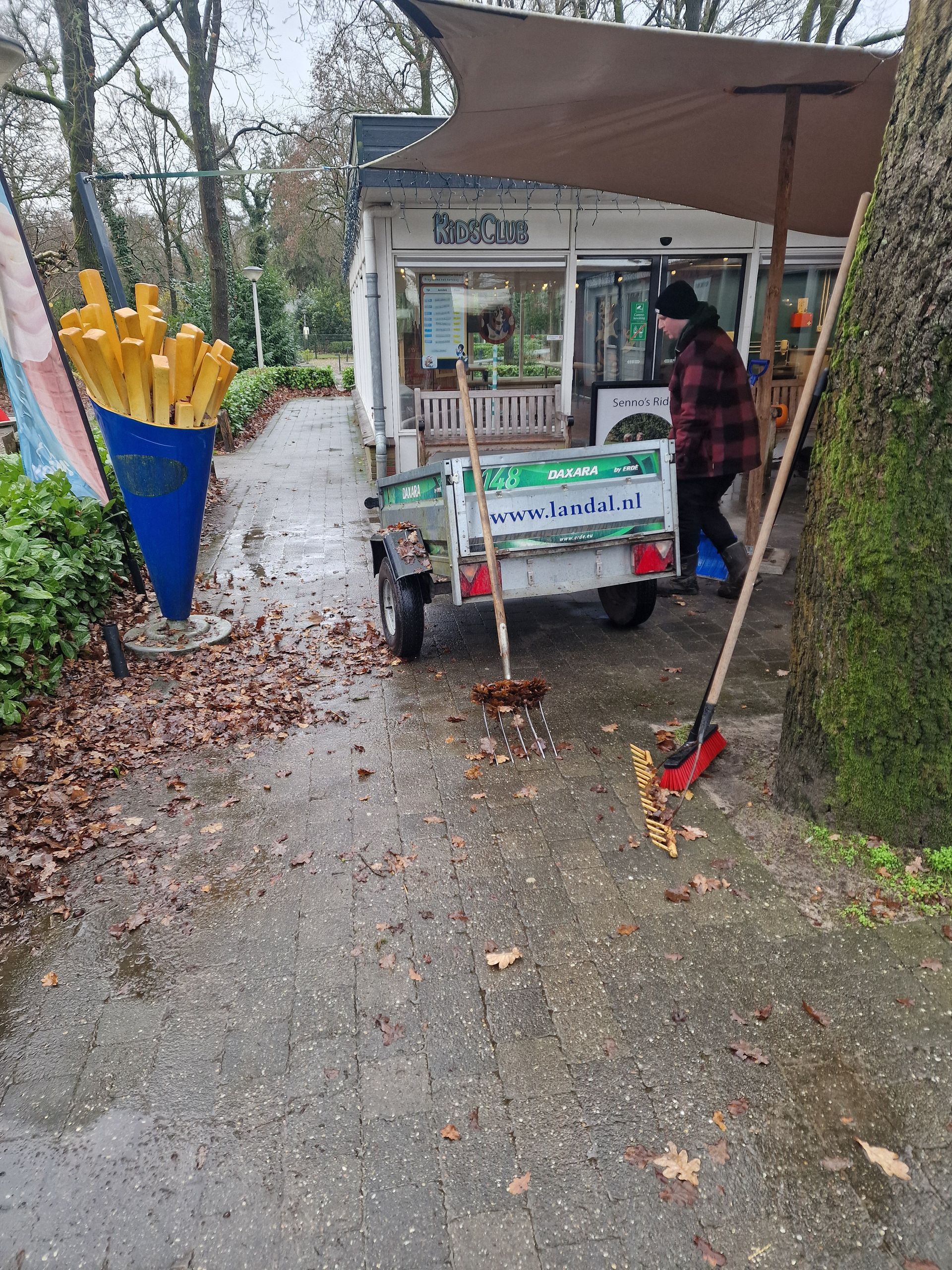 Iemand veegt bladeren op bij een eetkraam met een display voor frietpuntjes en een kleine aanhanger.