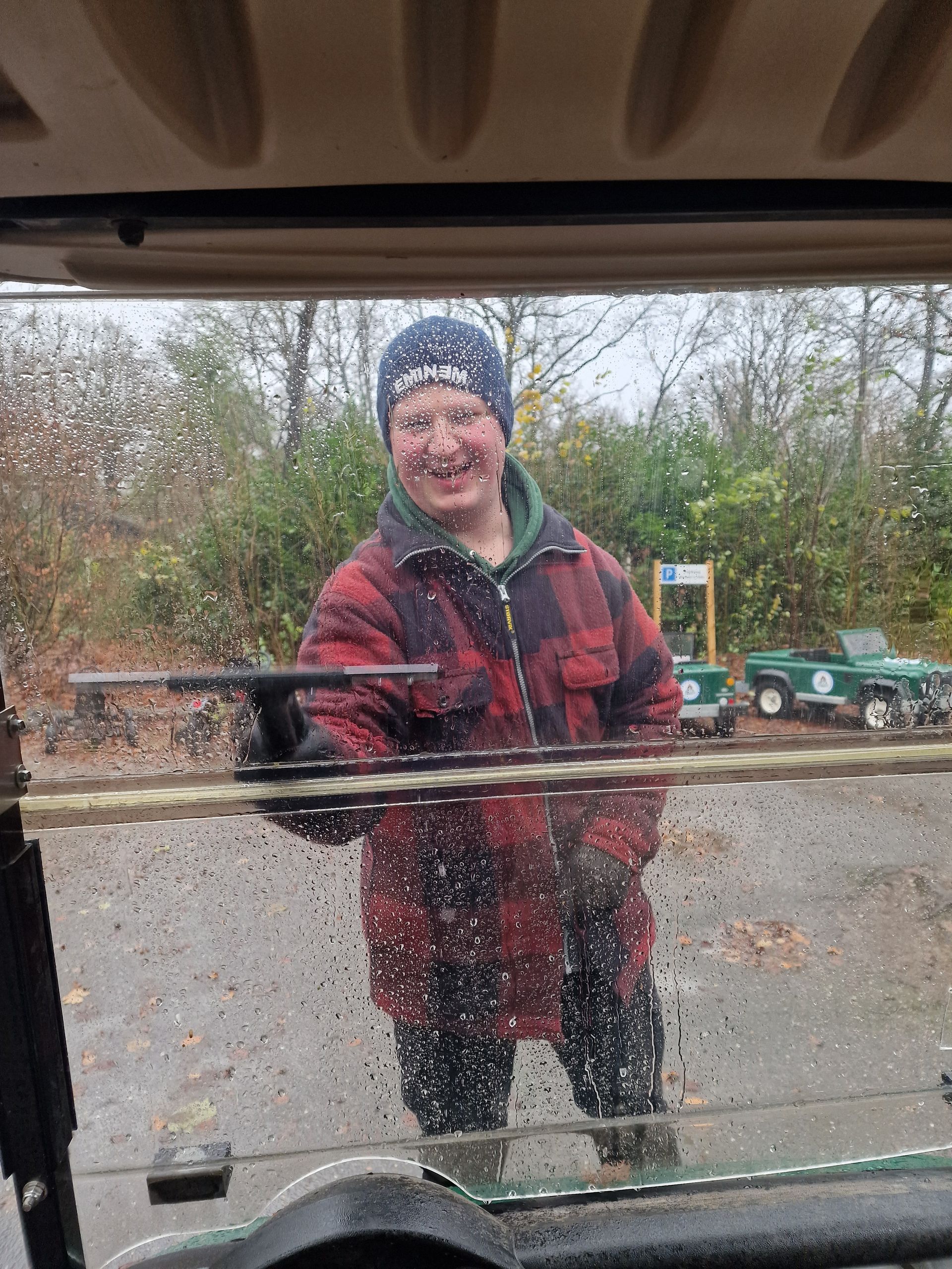 Een persoon in een rood geruit jasje lacht terwijl hij op een regenachtige dag de voorruit van een golfkarretje schoonveegt.