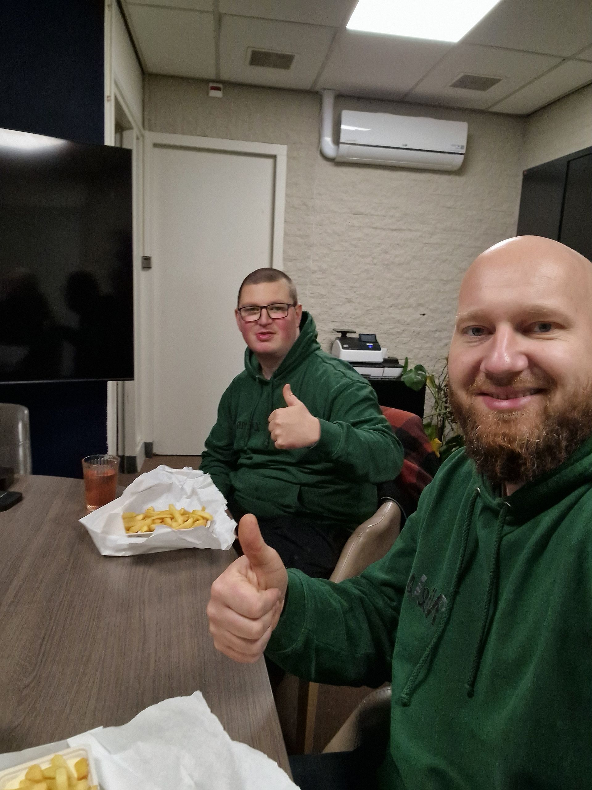 Twee mensen in groene hoodies steken hun duim omhoog terwijl ze binnen aan een tafel friet eten.