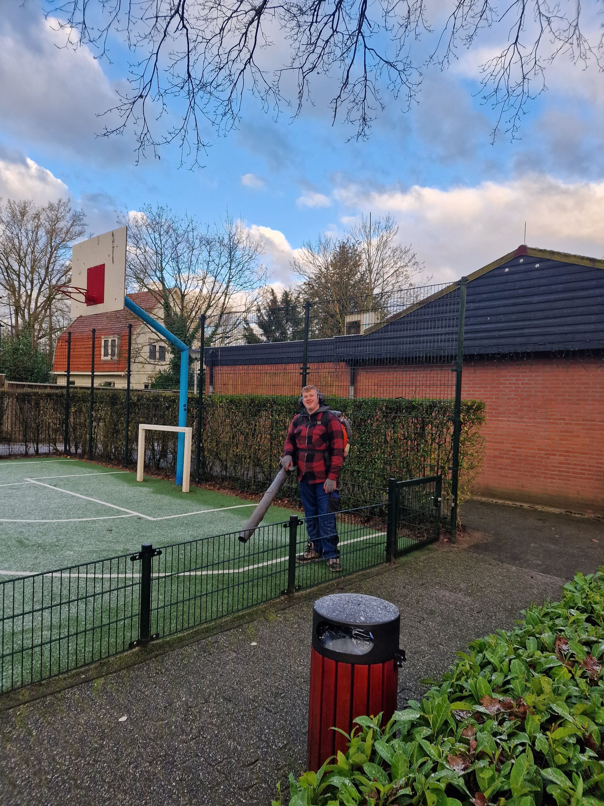 Man in geruit shirt gebruikt bladblazer op een groen speelveld; basketbalring en gebouw op de achtergrond.