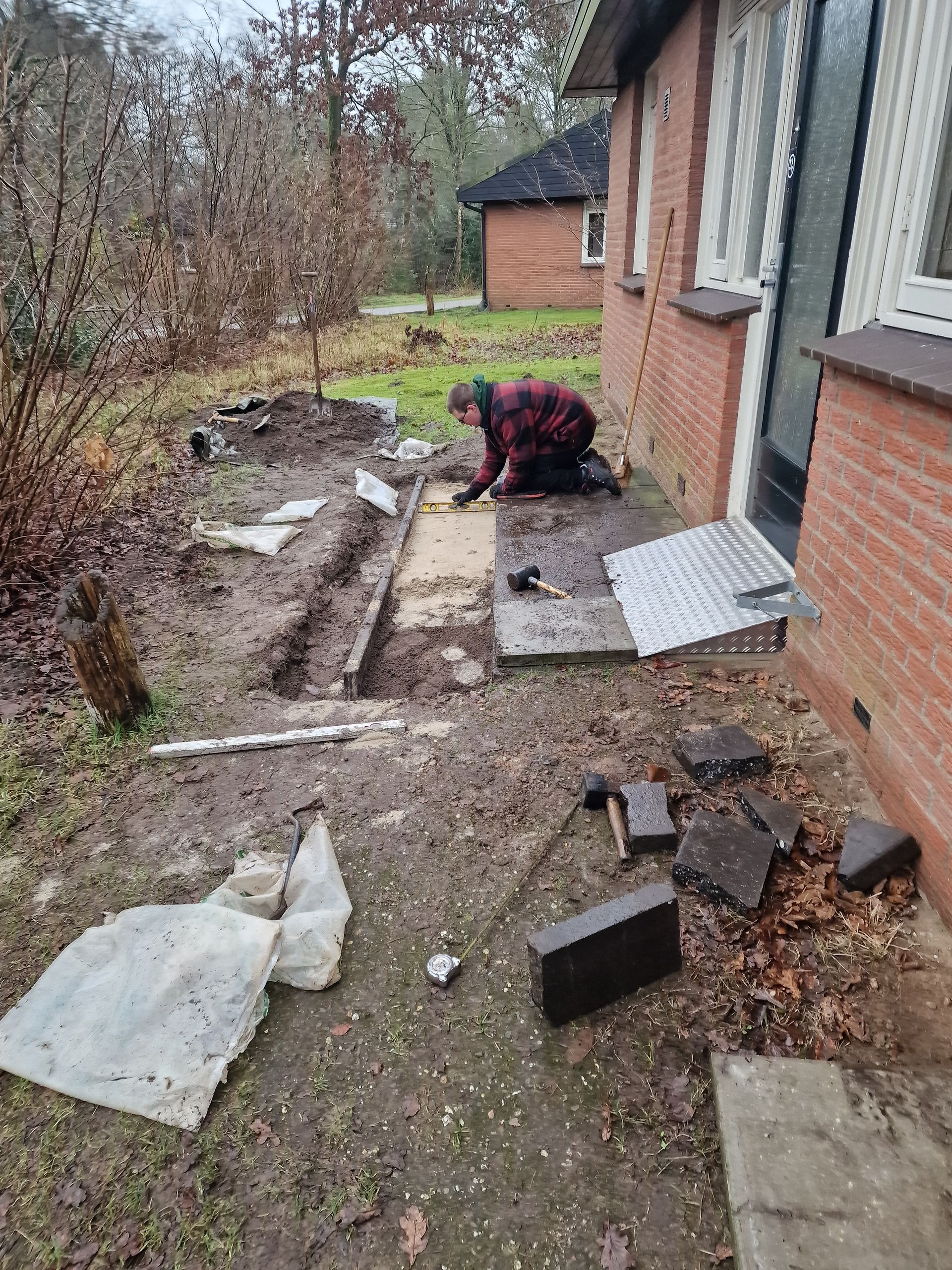 Een persoon knielt en werkt aan een stenen terras naast een huis. Gereedschap en materialen liggen op de grond.