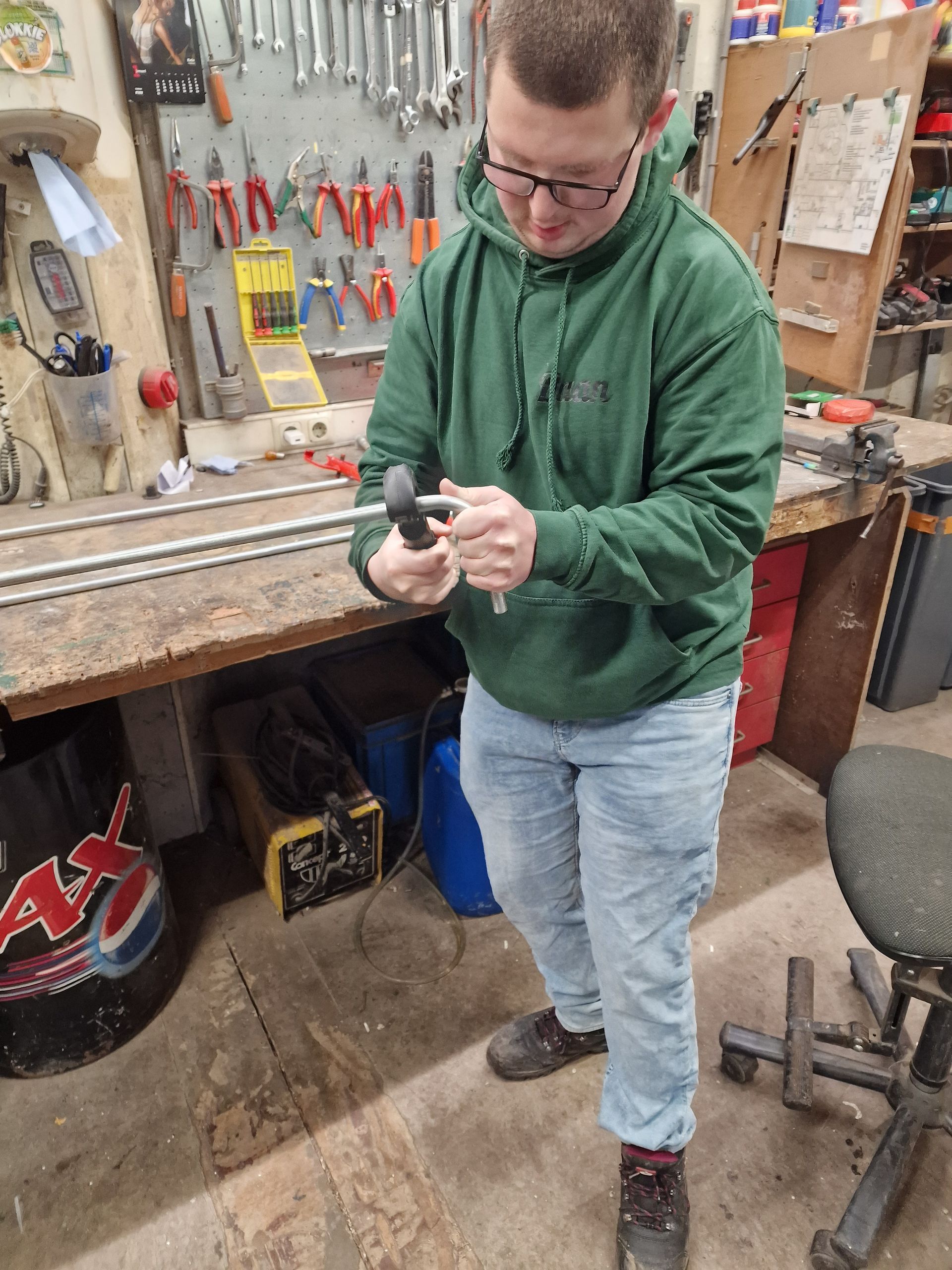 Man in groene hoodie en bril aan het werk met gereedschap in een werkplaats.
