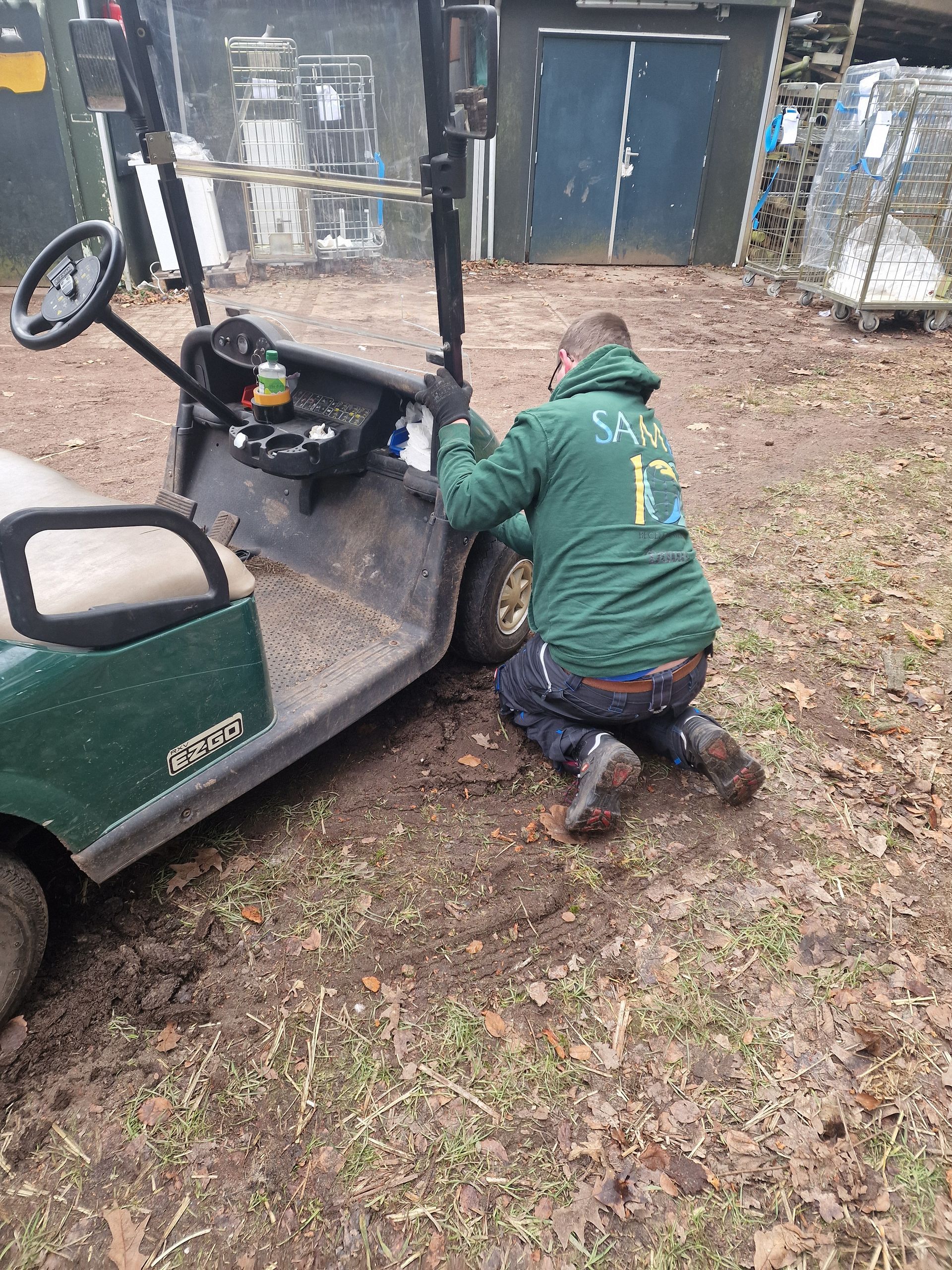 Een persoon knielt naast een groene golfkar, mogelijk om deze buiten te repareren.