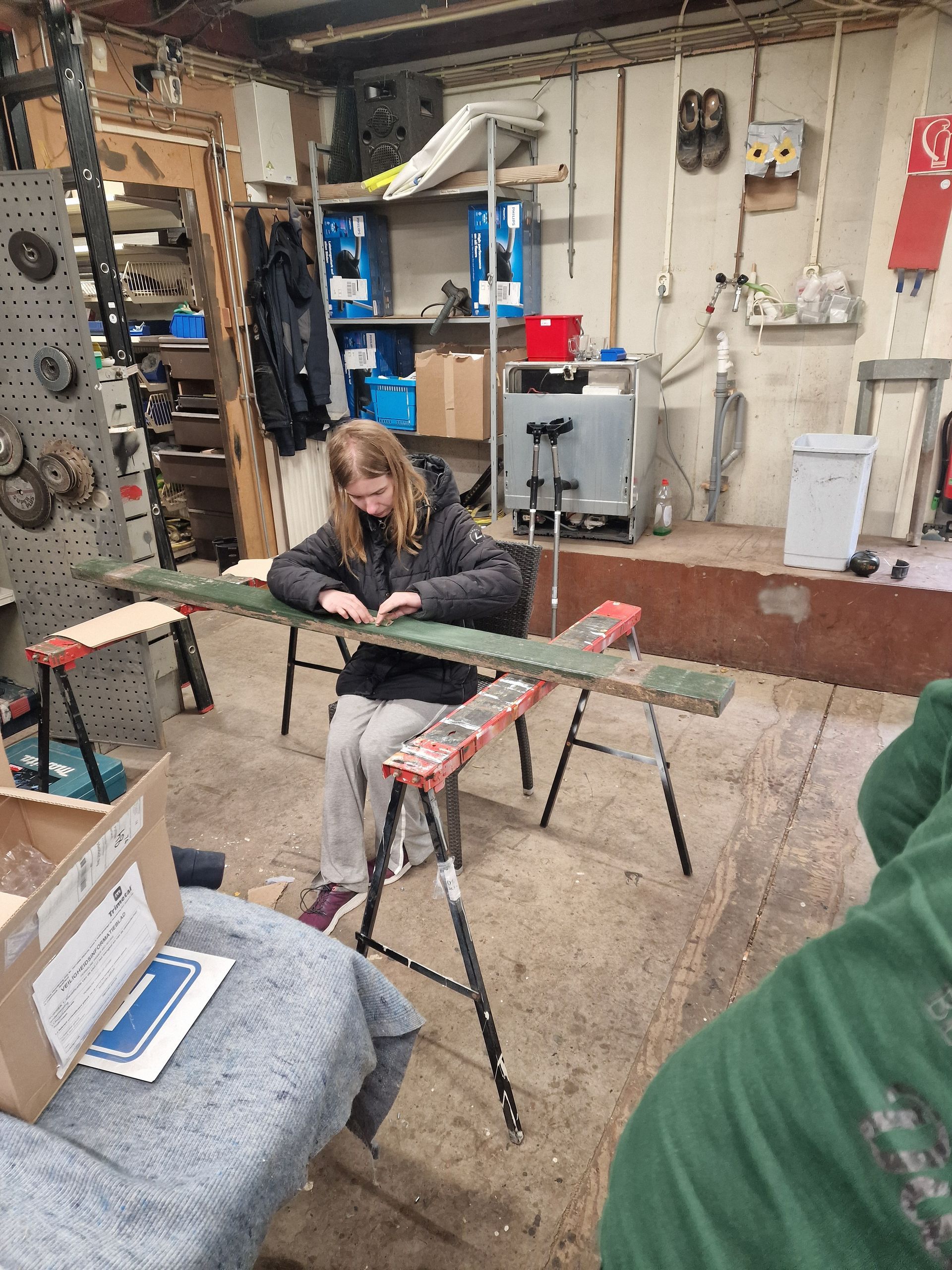 Een persoon zit en leunt tegen een plank die op schragen rust in een werkplaats.