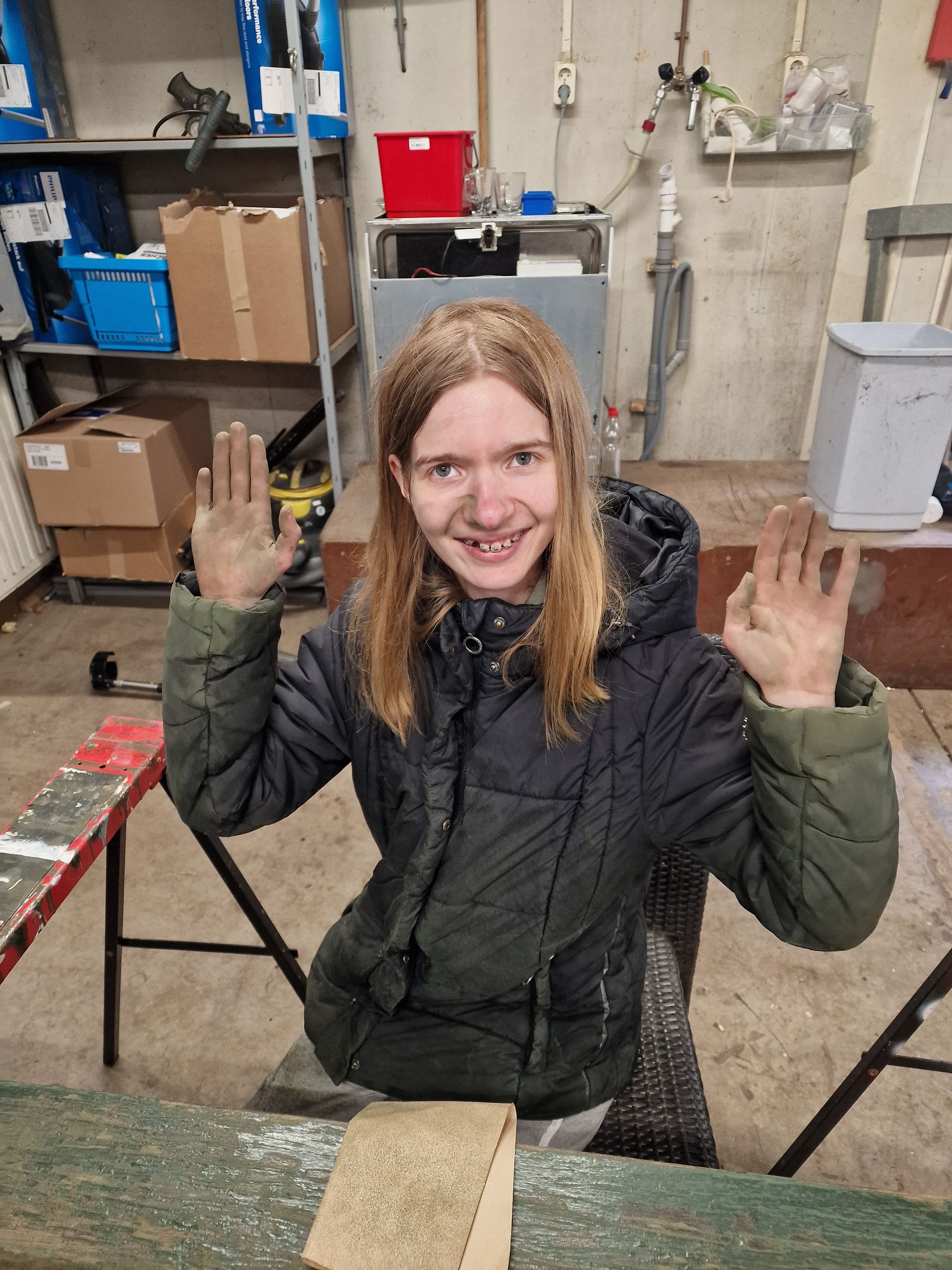 Een persoon met blond haar, een zwarte jas aan, steekt zijn handen omhoog en lacht in een werkplaats.