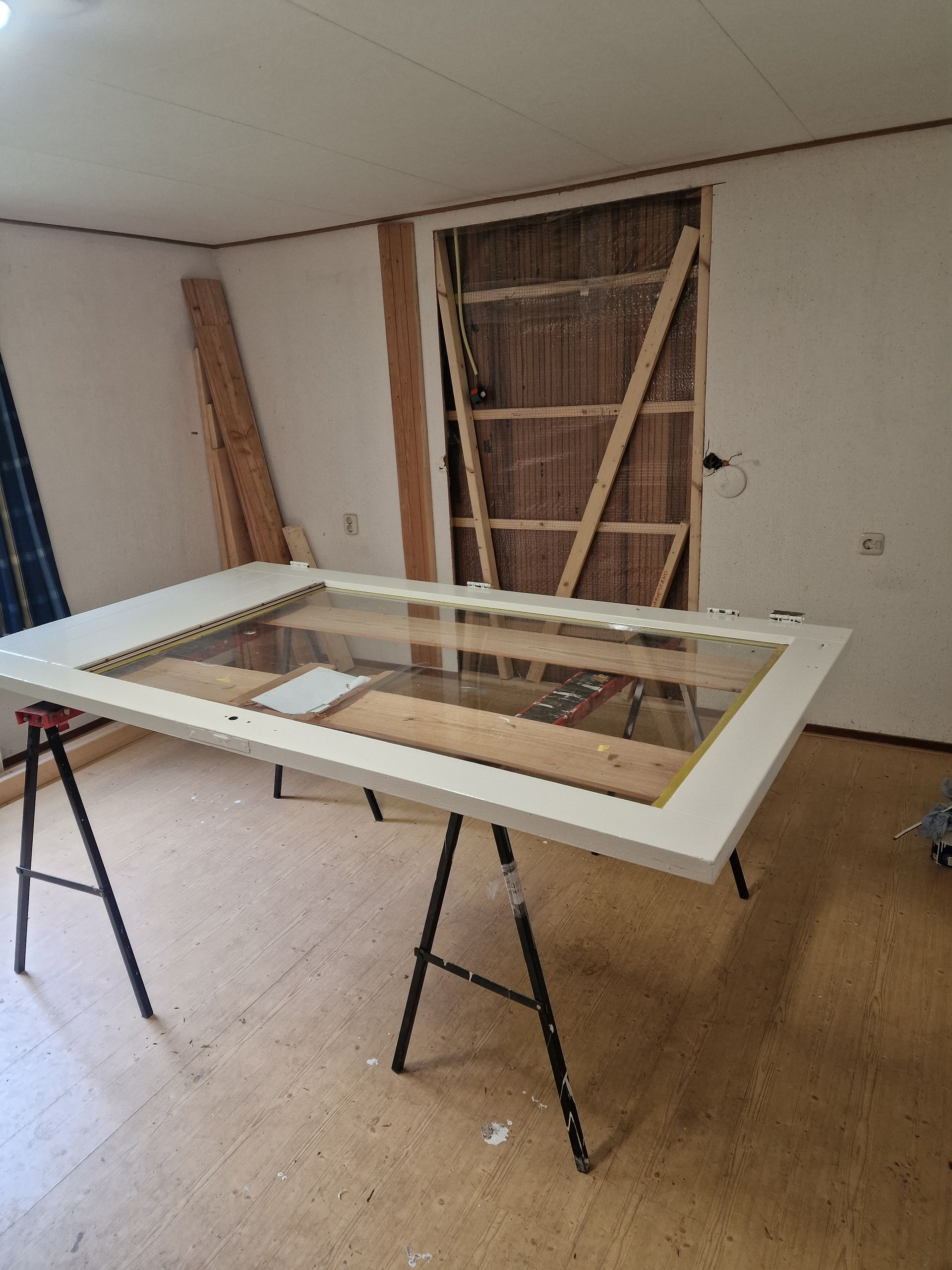 Grote glazen ruit in een wit frame op een schraag; de kamer is in aanbouw.
