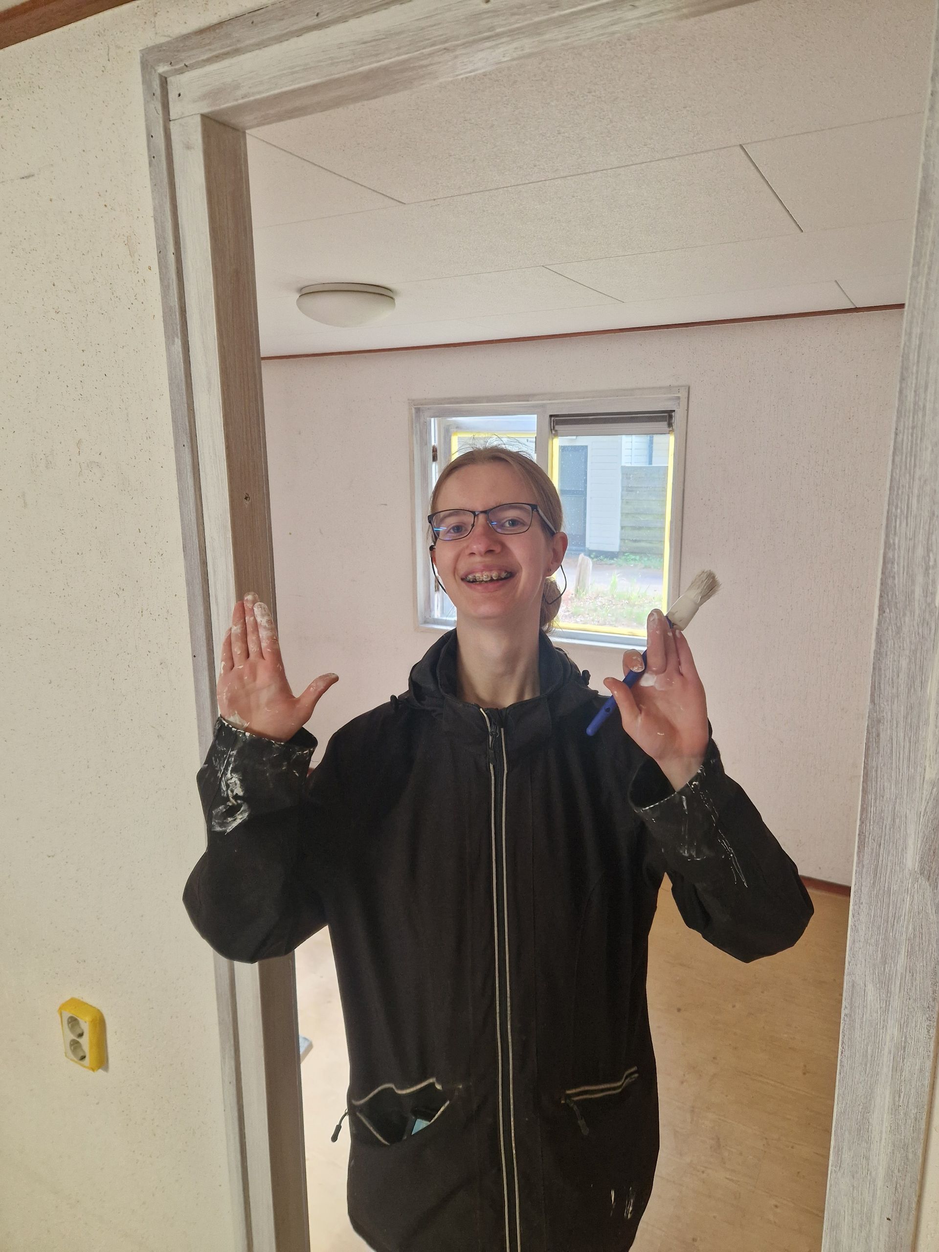 Een vrouw met een bril en een zwarte jas staat in de deuropening, gebaart en glimlacht. Interieurbeeld van een lege kamer.