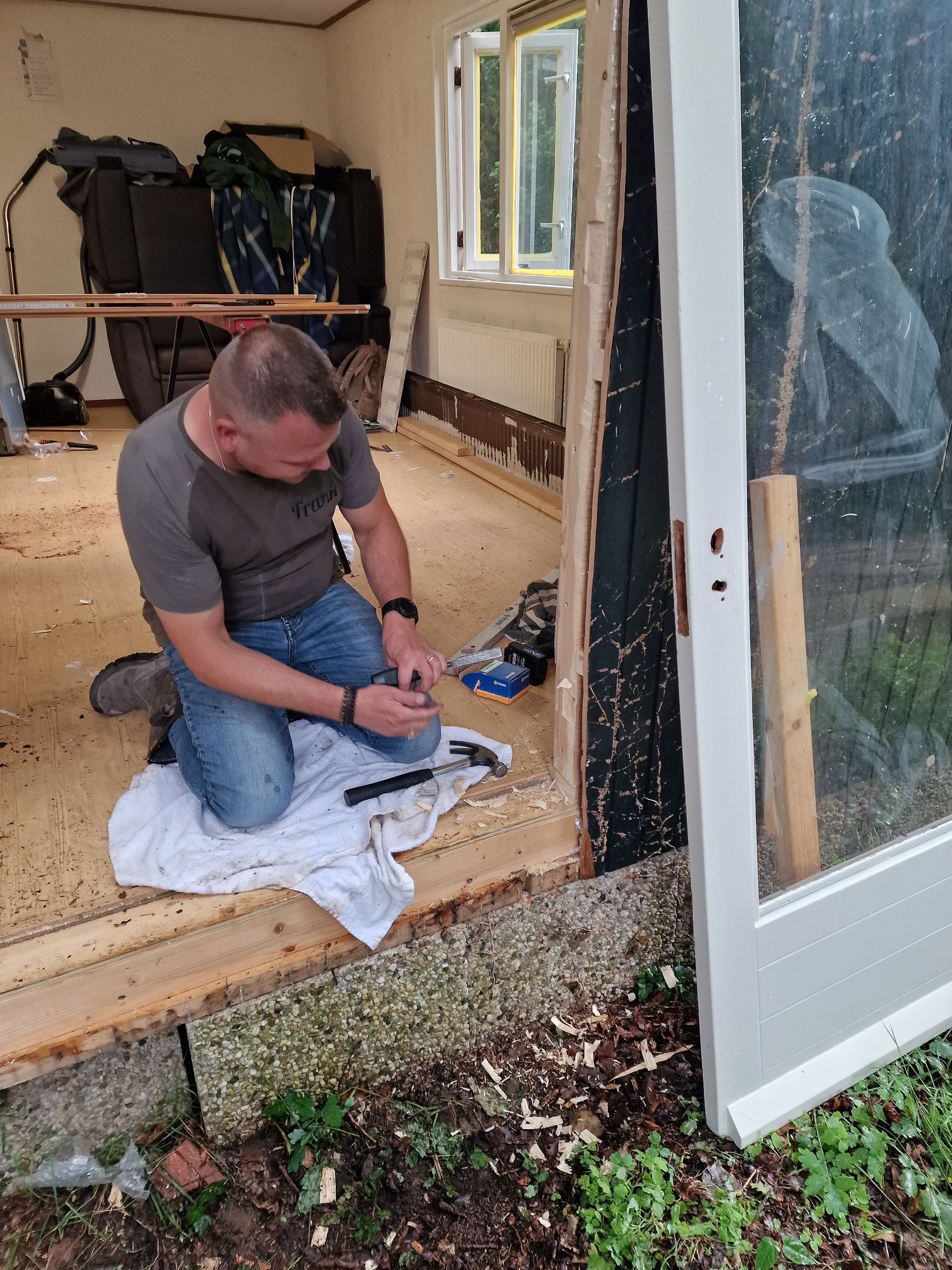 Een man knielt en bewerkt met een beitel een houten deurkozijn. Zonlicht stroomt naar binnen. Zaagsel ligt op de vloer.