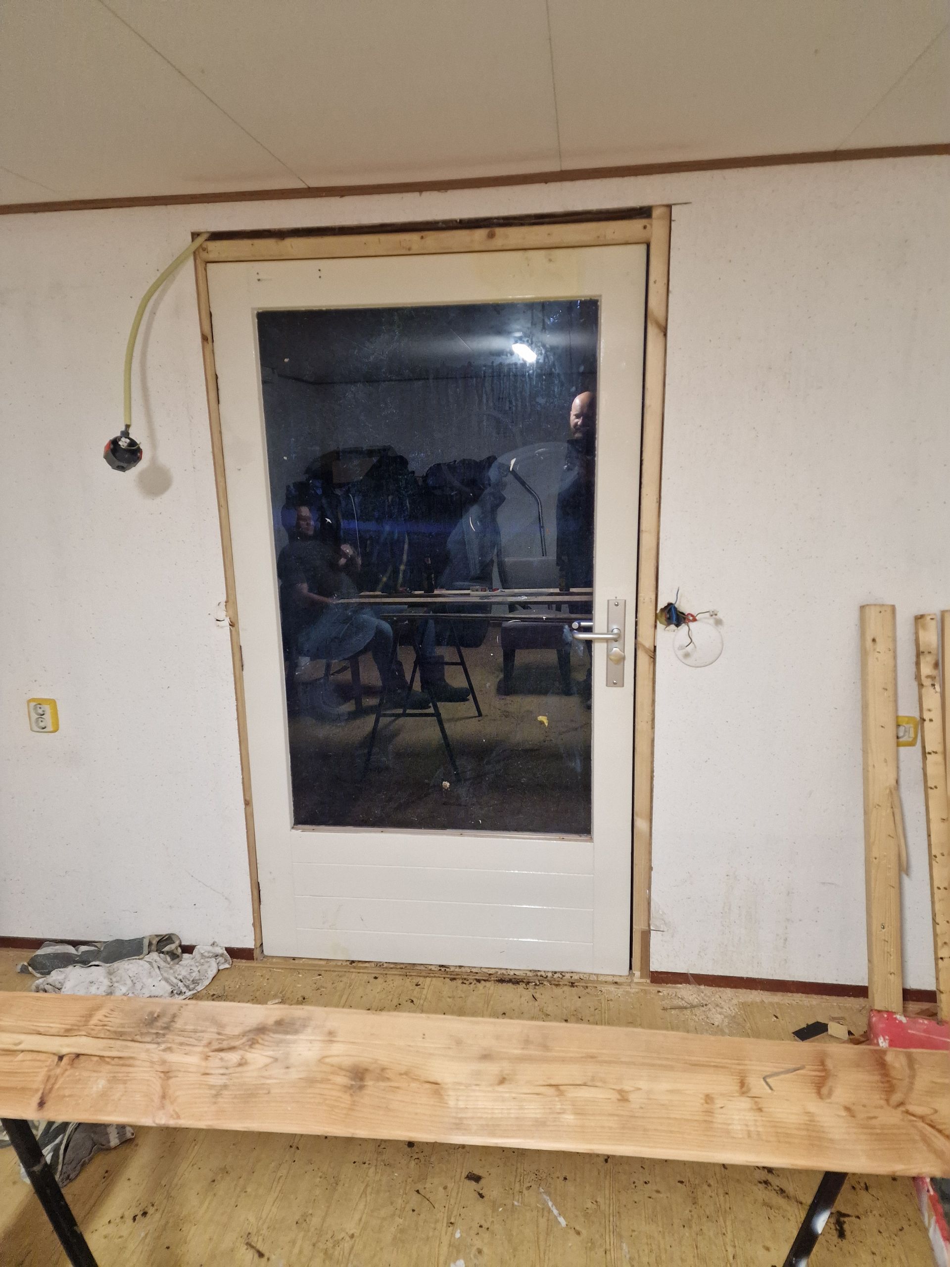 Deur met houten frame, binnenkant, onafgewerkt. De deur is wit, met een glazen paneel waarin de reflectie zichtbaar is.