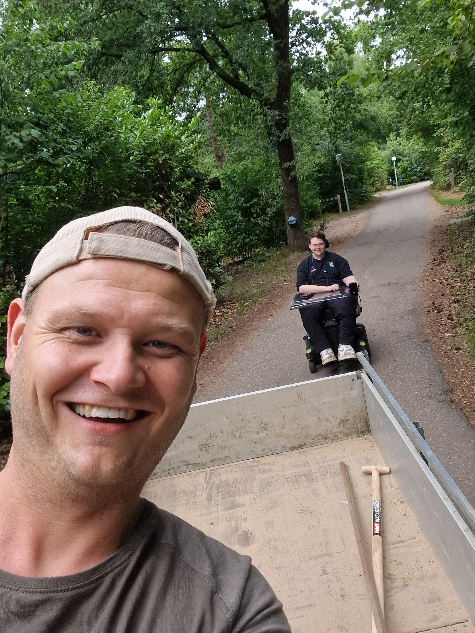 Een lachende man maakt een selfie met een persoon in een rolstoel op een pad. Buitenomgeving met bomen en een weg.