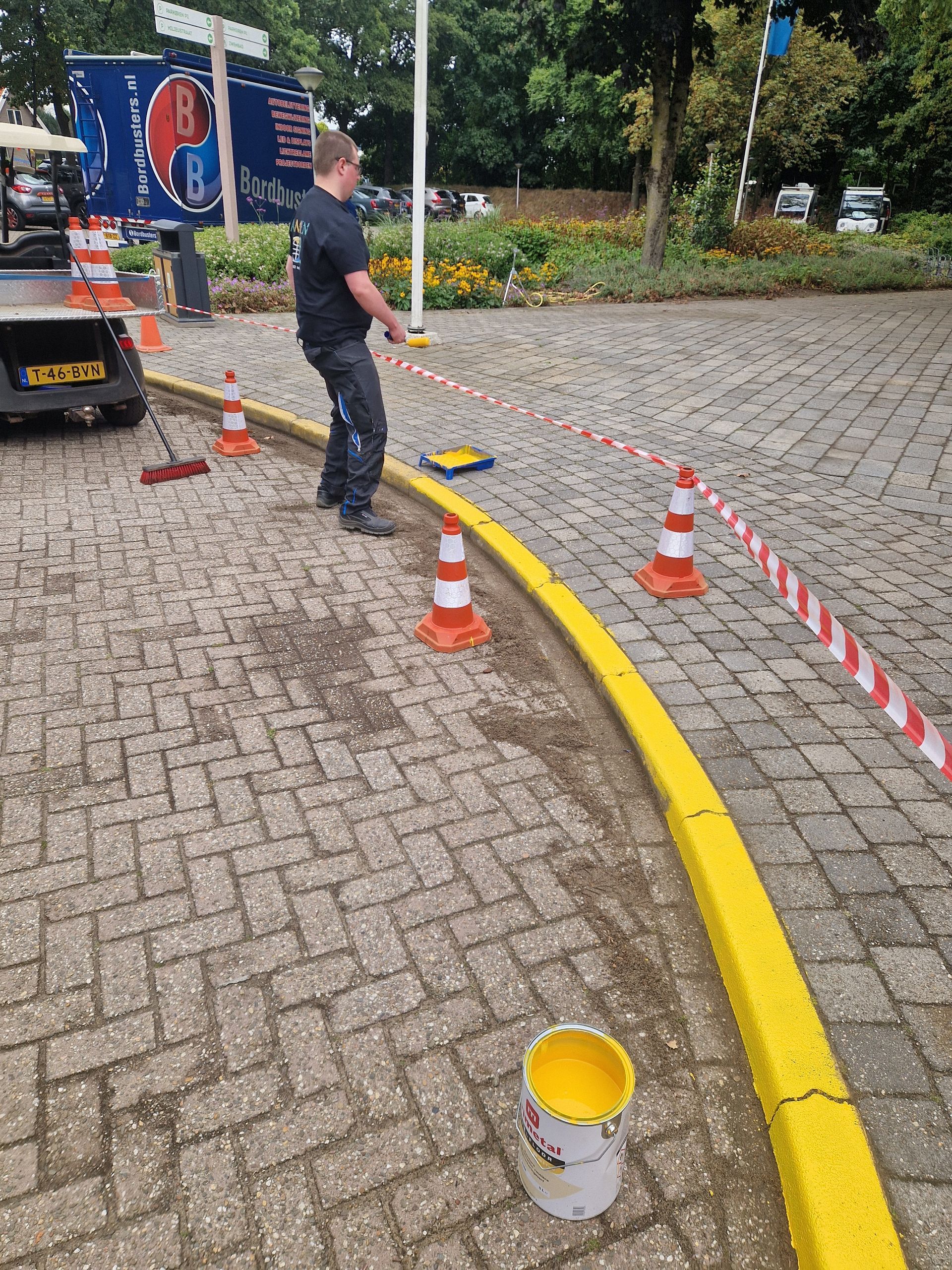 Iemand schildert een gele stoeprand op bakstenen bestrating, waarbij kegels en afzetlint voor de veiligheid worden gebruikt.