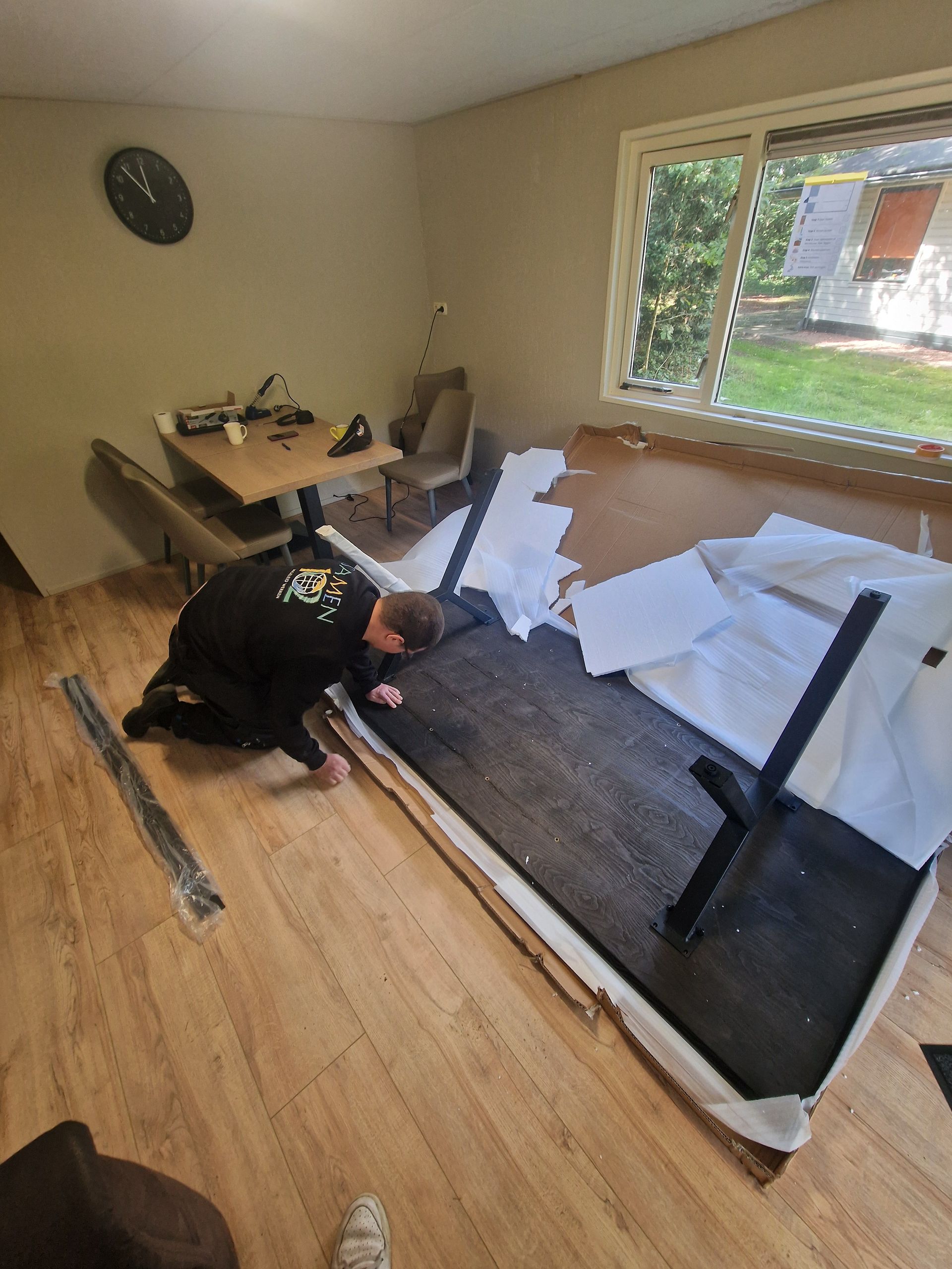 Een persoon knielt naast een beschadigde bank in een kamer met een tafel, stoelen en een raam.