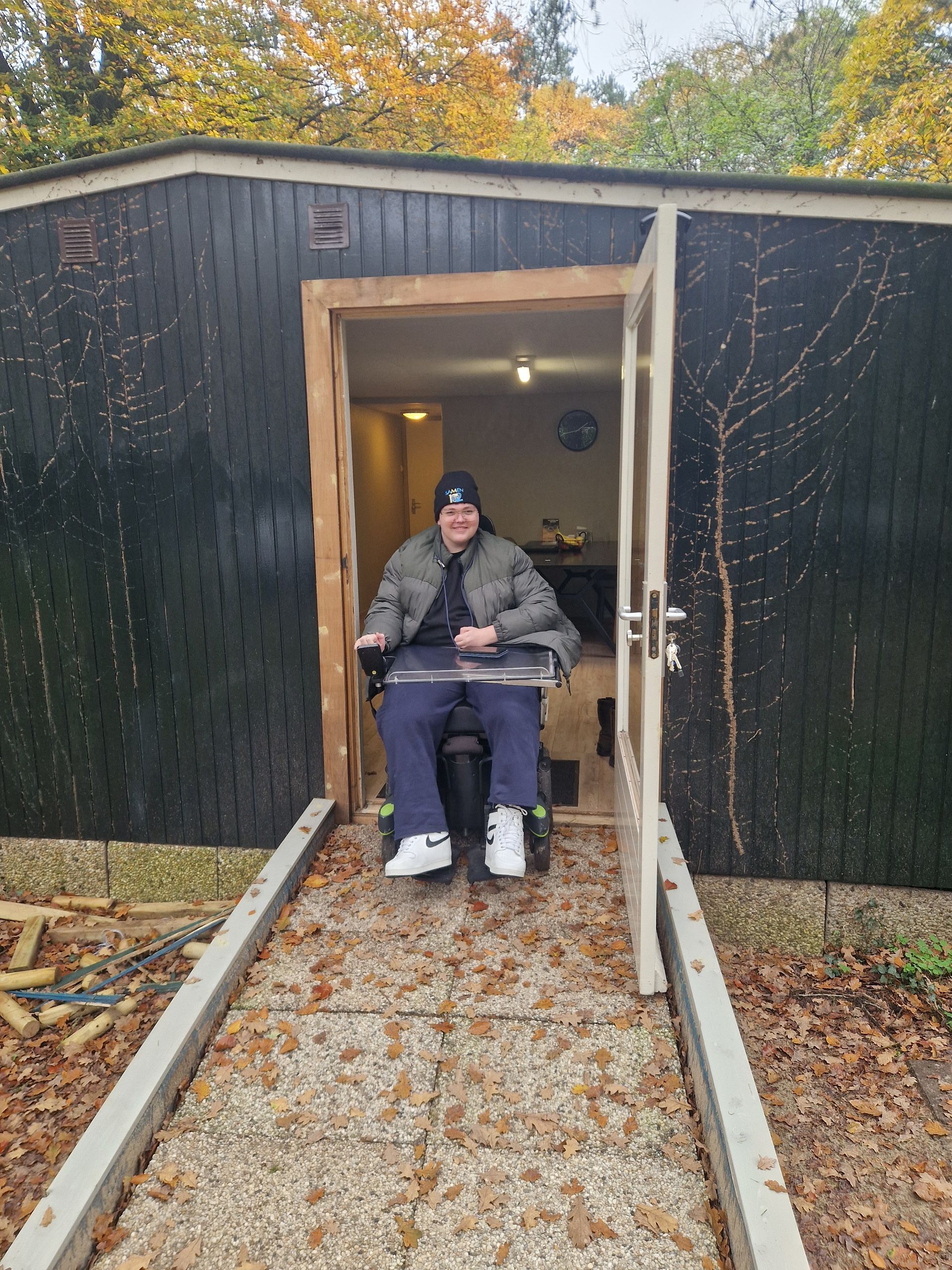 Een persoon in een rolstoel verlaat een donkergekleurde cabine met een hellingbaan. De deuropening heeft een houten kozijn.