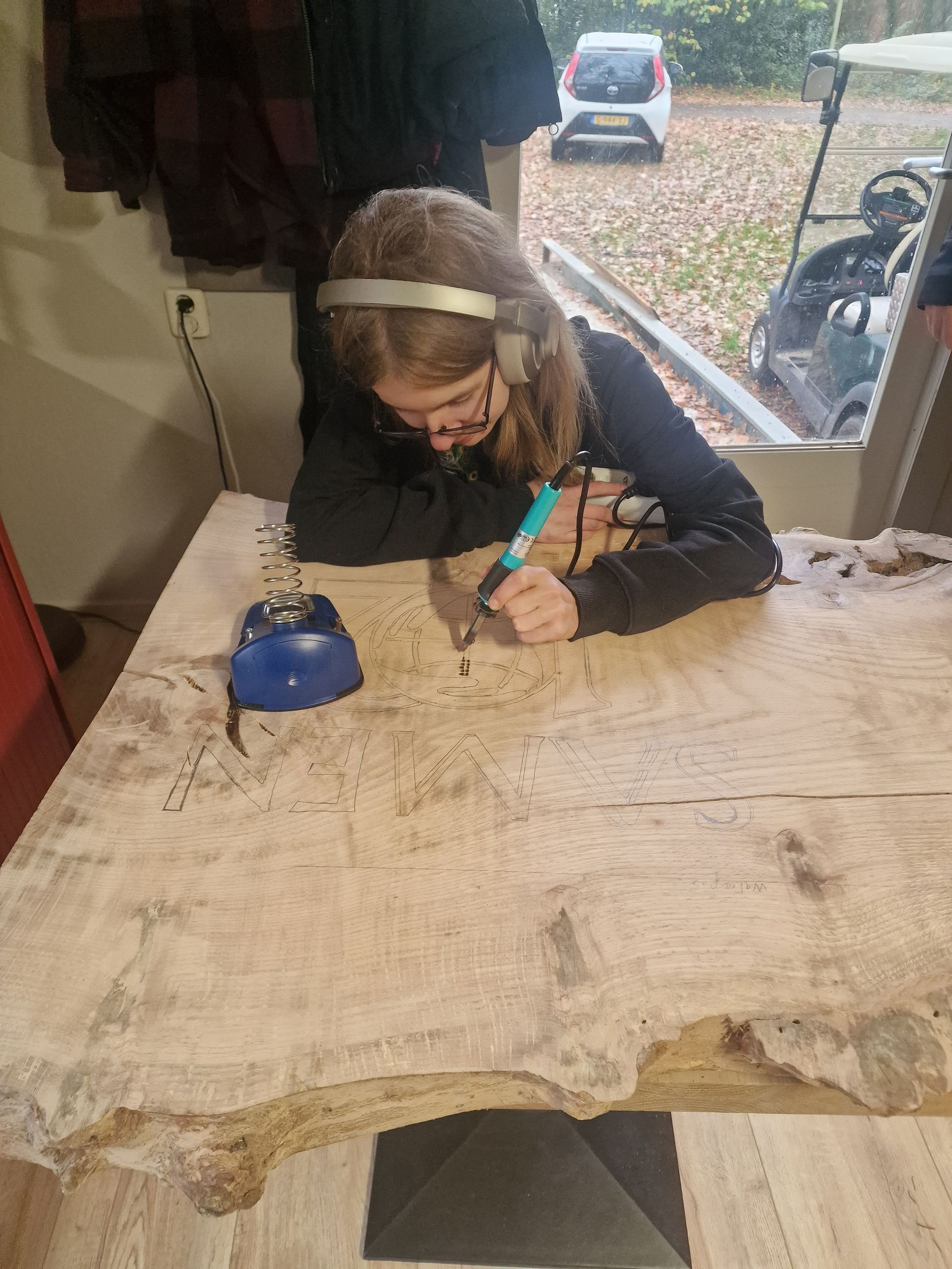 Een persoon tekent op een houten tafel, draagt ​​een bril en een haarband. Een blauw strijkijzer staat ernaast.