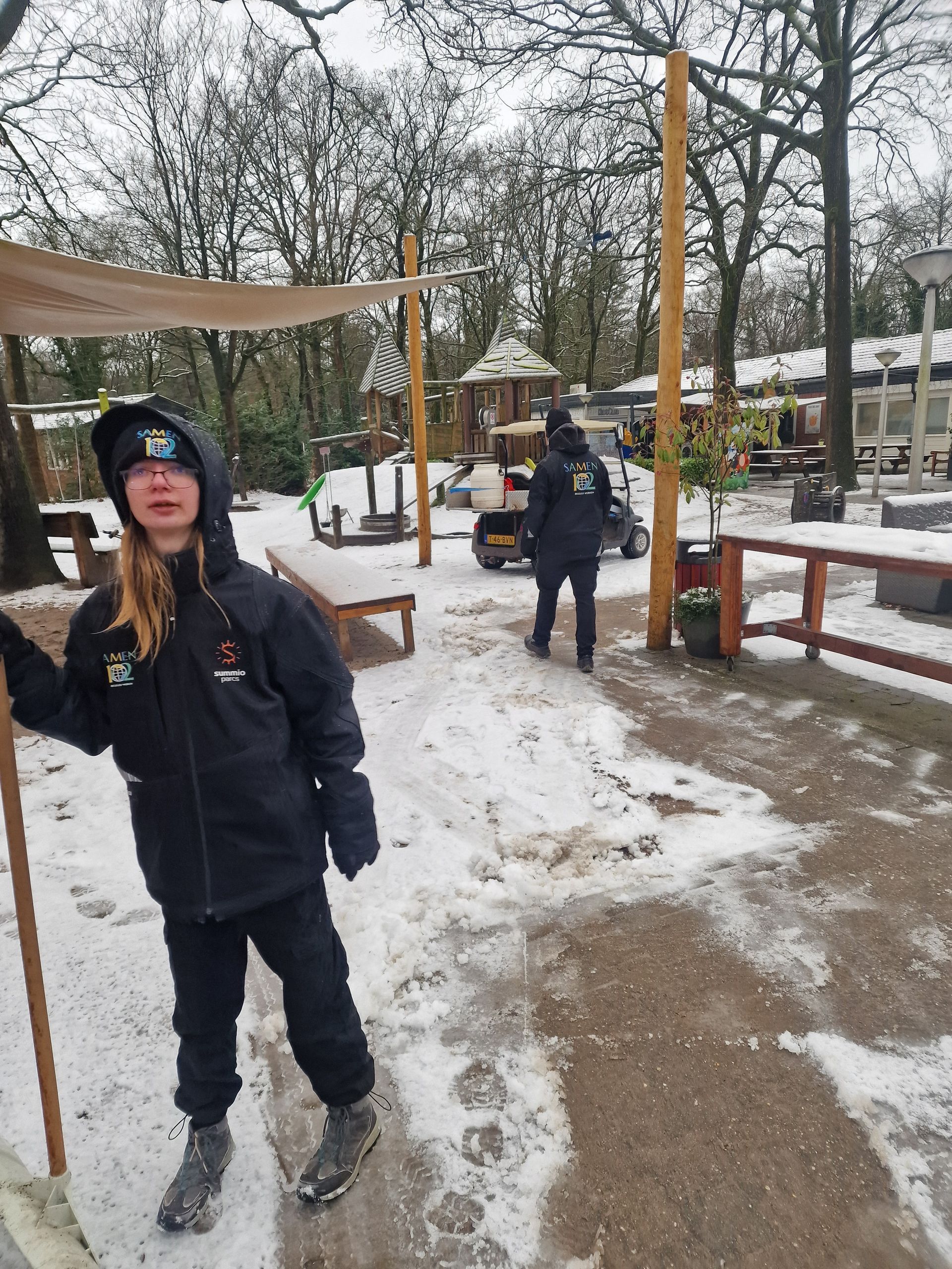 Een persoon in zwarte bovenkleding staat met een staf in een besneeuwd gebied; een andere persoon loopt op de achtergrond.
