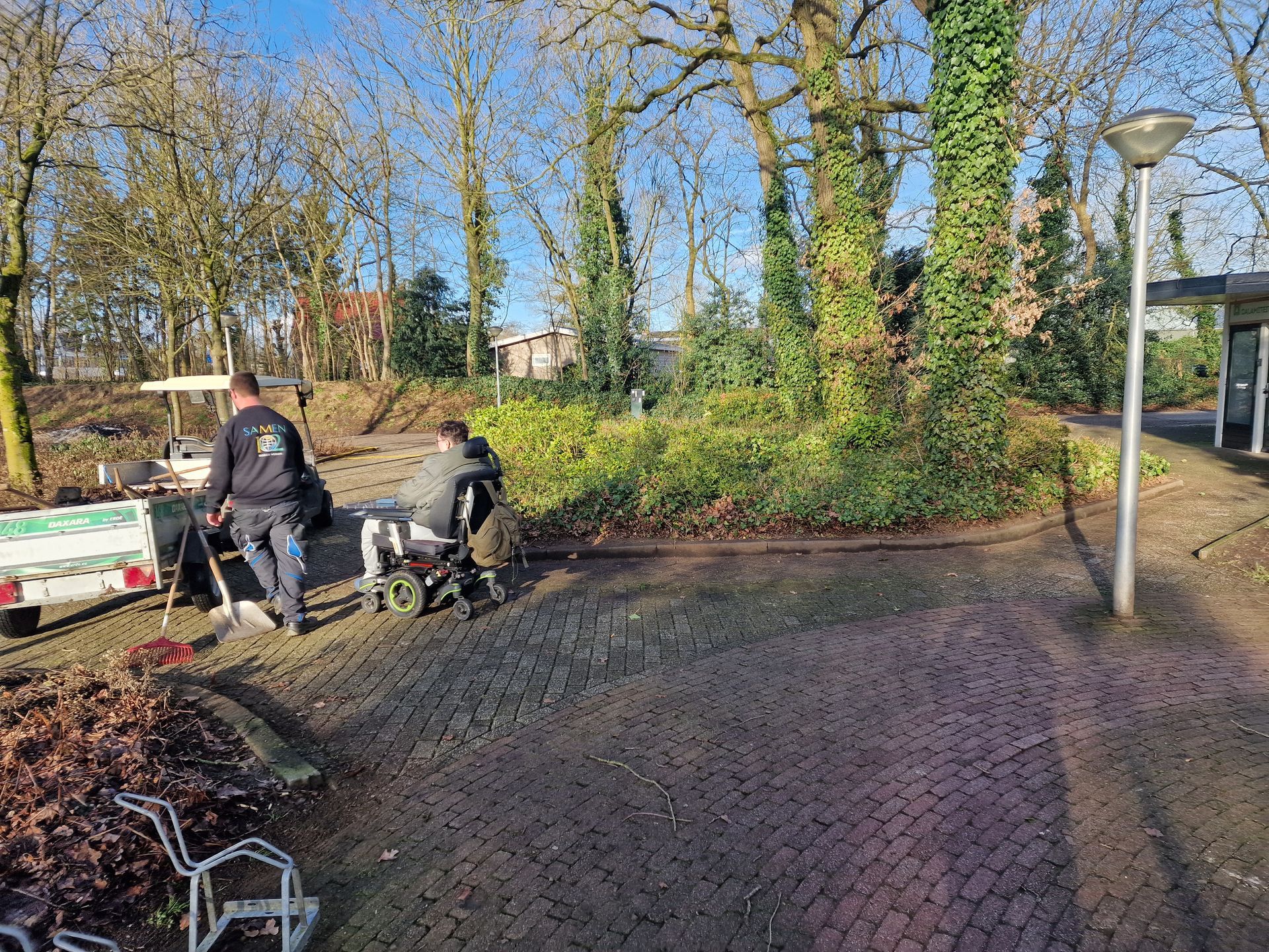 Twee arbeiders staan ​​bij een aanhanger met apparatuur, omringd door bomen en een bakstenen pad.