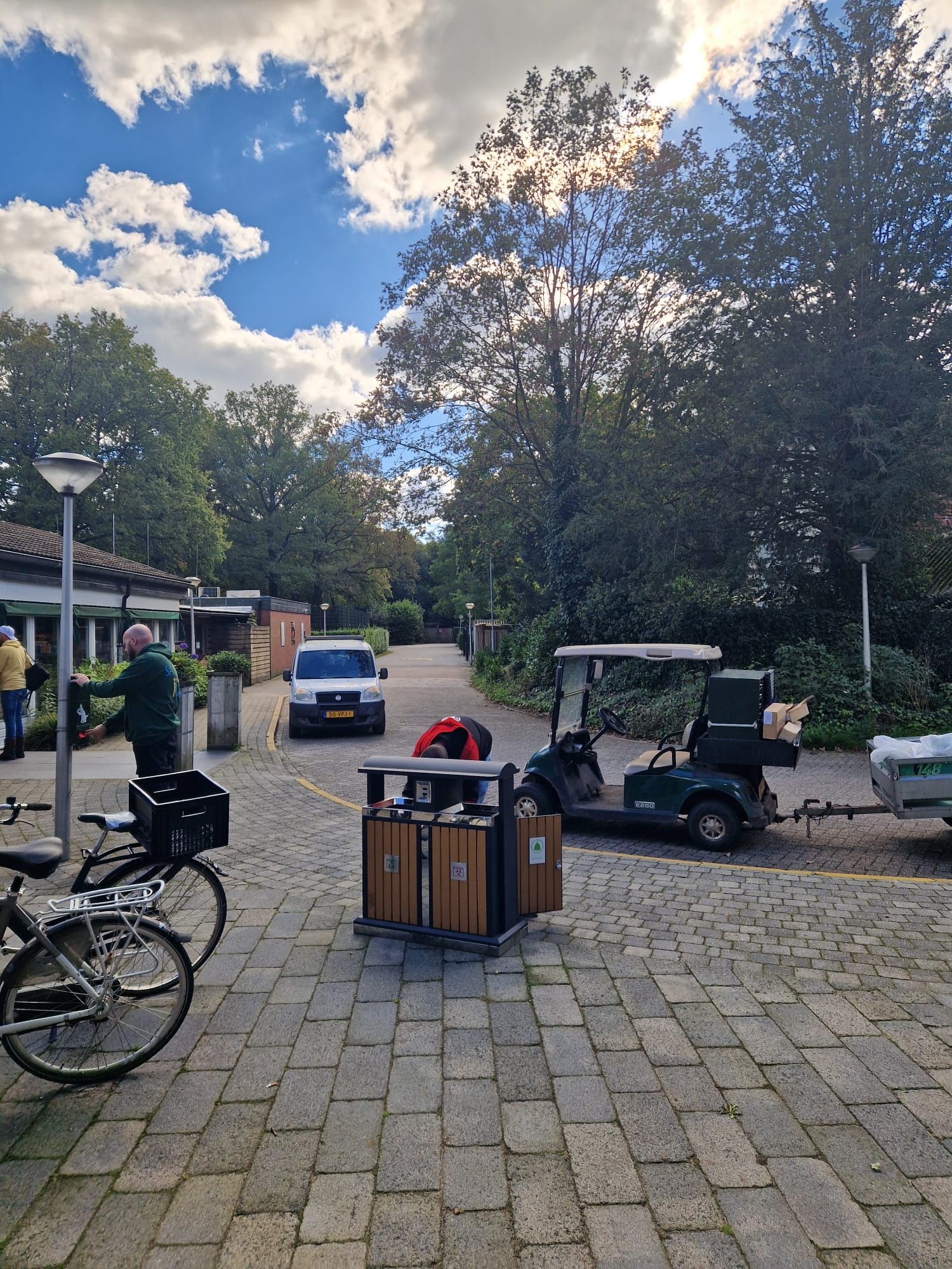 Verharde weg met bomen, een geparkeerde golfkar, vuilnisbakken en een persoon in de buurt. Zonnige dag.