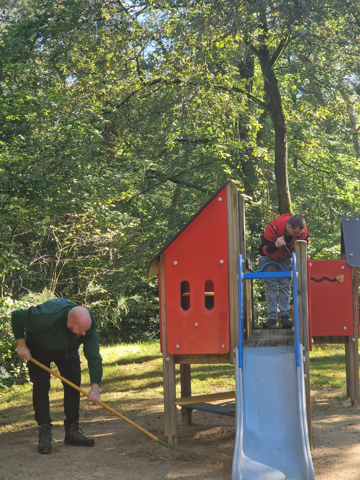 Twee mensen maken speeltoestellen in een park schoon, de een veegt, de ander inspecteert.