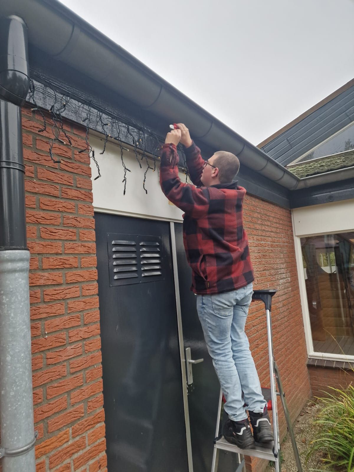 Een persoon staat op een ladder en hangt lichtsnoeren aan de dakgoot van een gebouw.