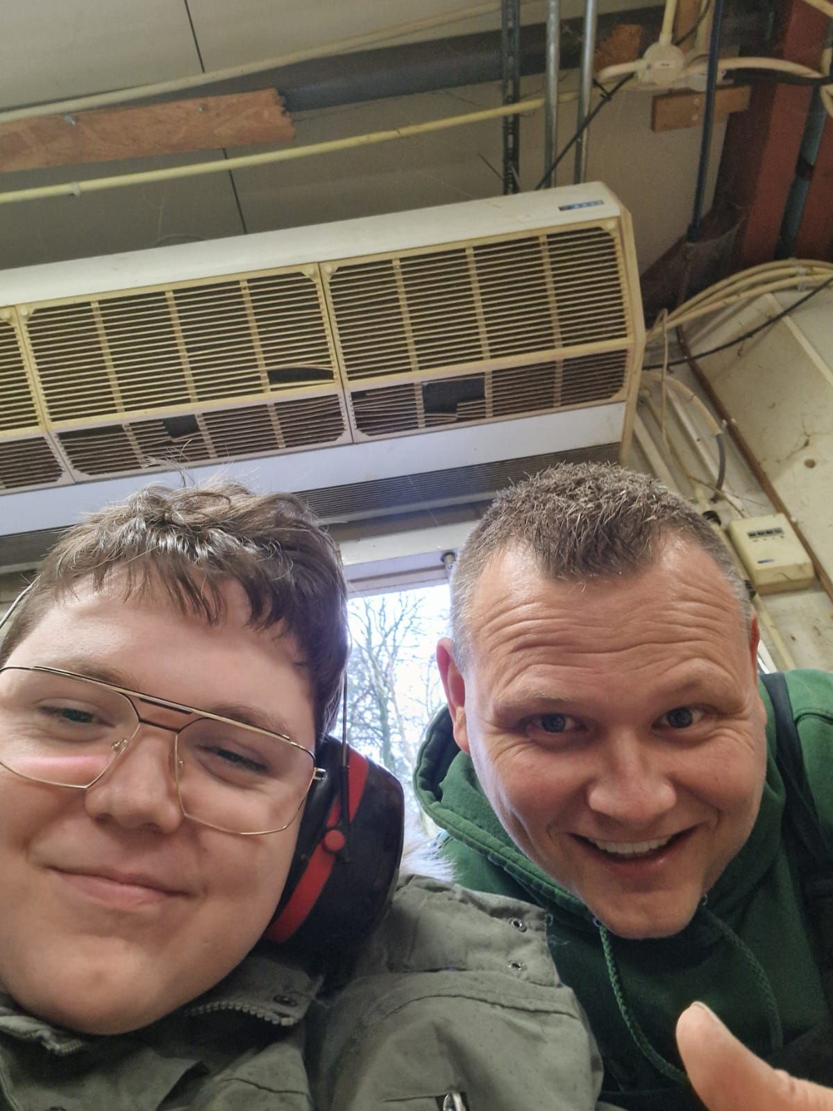Twee mensen glimlachen, een met een bril en de ander met een duim omhoog, binnen bij een airconditioningunit.