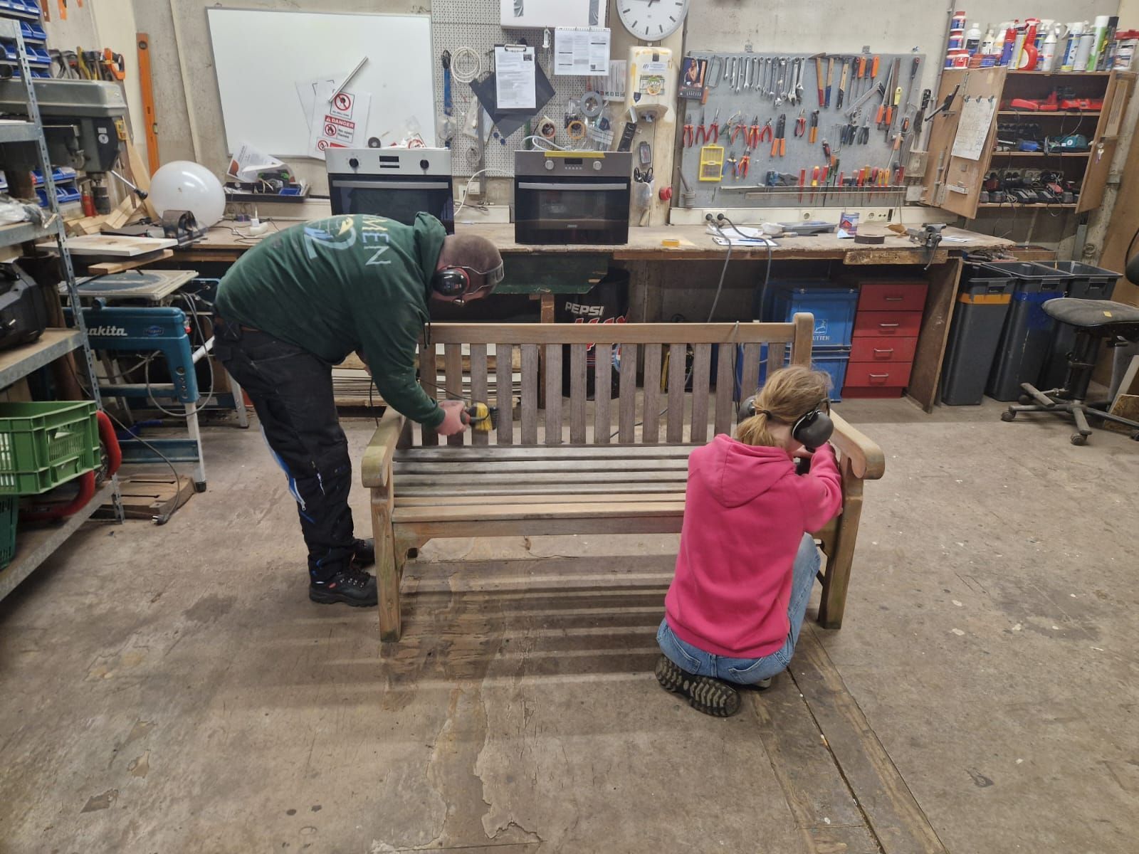 Twee mensen repareren een houten bank in een werkplaats; de een schuurt, de ander gebruikt gereedschap.