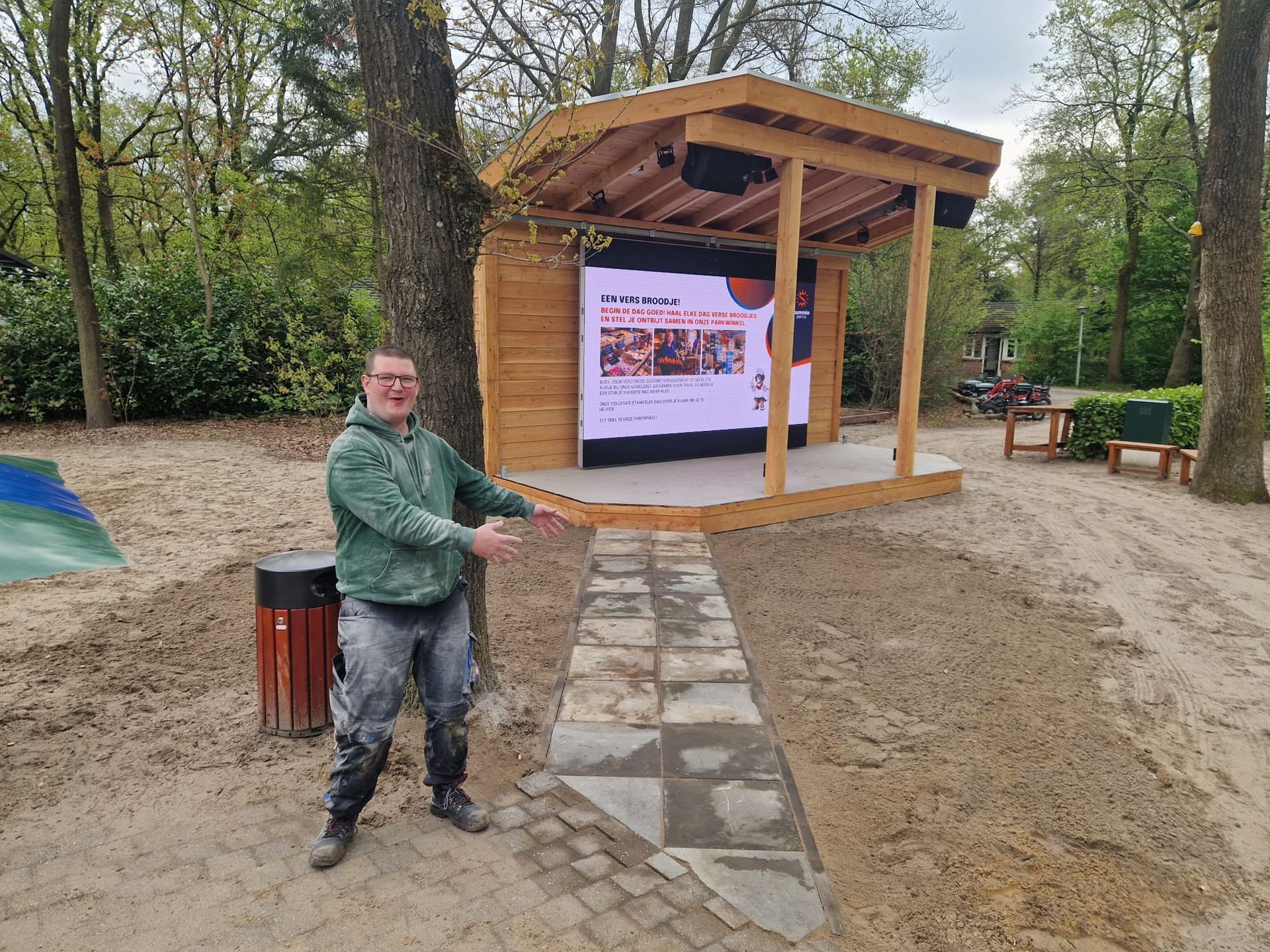 Een man wijst naar een houten podium met een groot scherm waarop een evenement wordt getoond. Buiten, in een park.