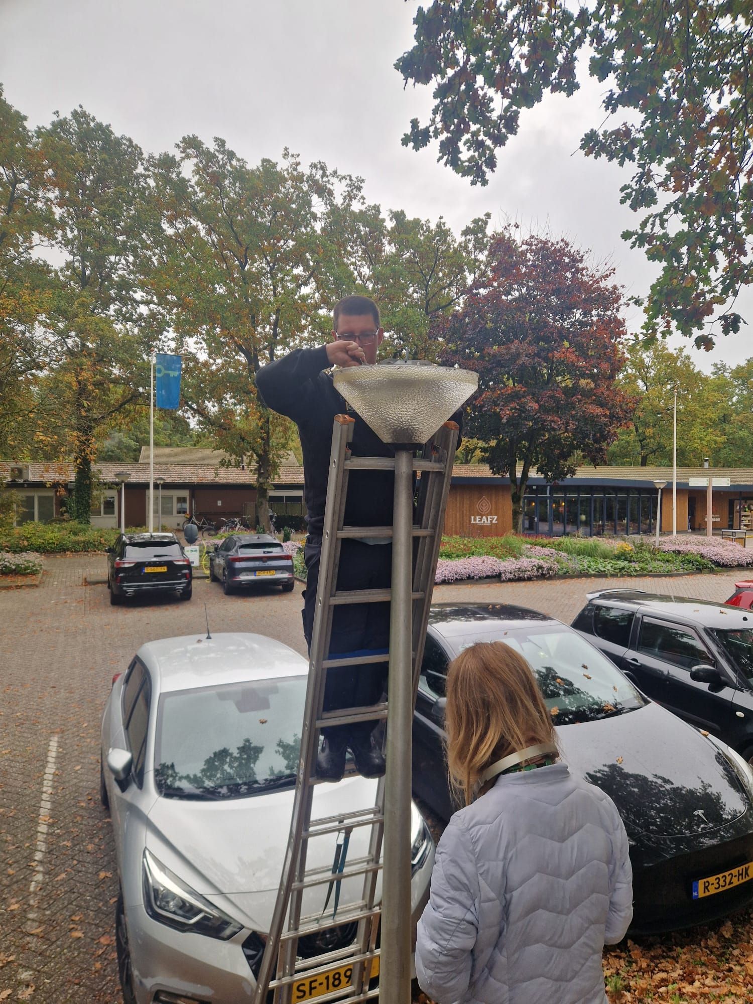 Een man staat op een ladder en repareert een straatlantaarn, geholpen door iemand anders. 