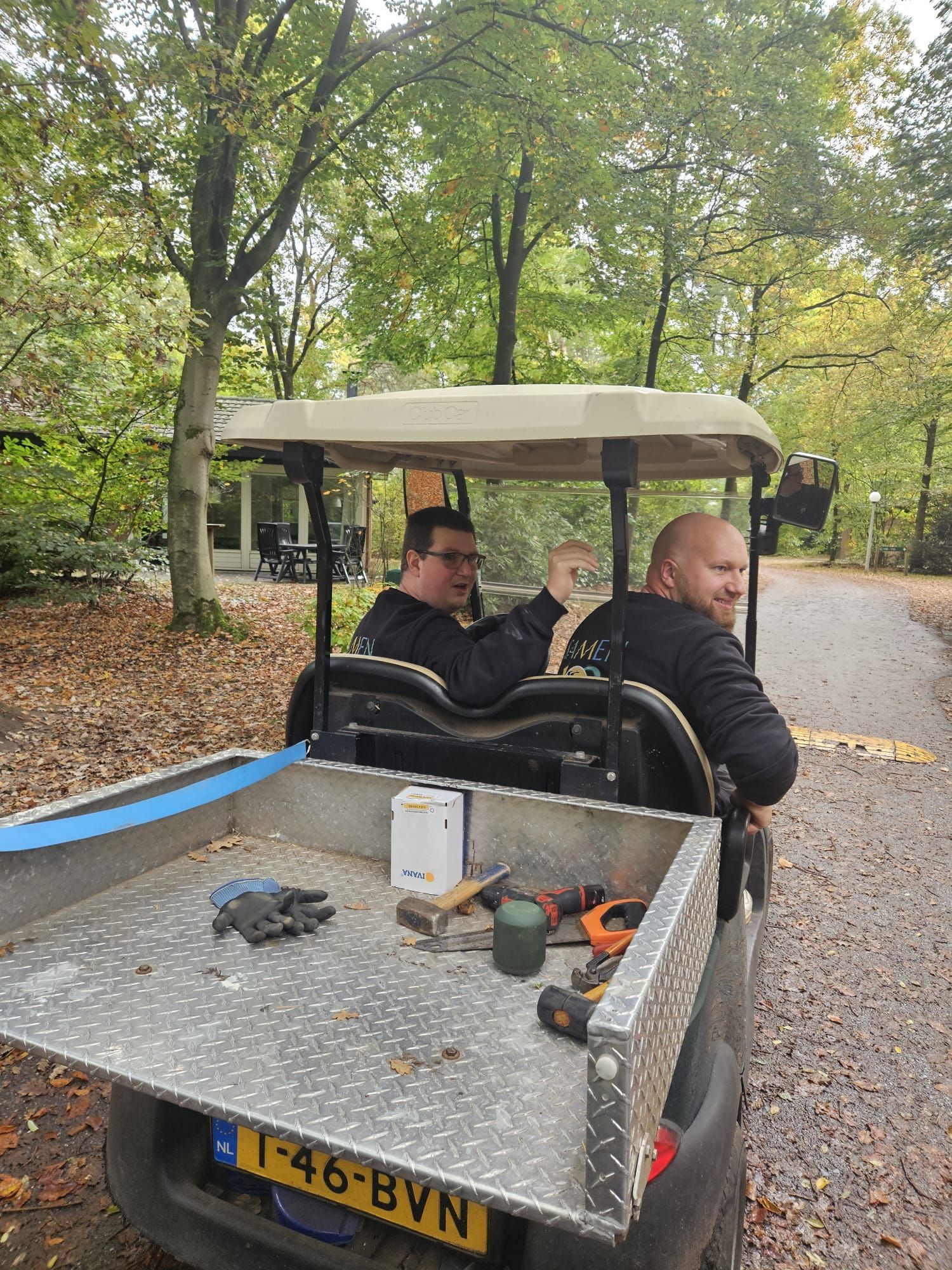 Twee mannen in een golfkarretje met gereedschap achterin, rijdend over een pad in een bosrijk gebied.