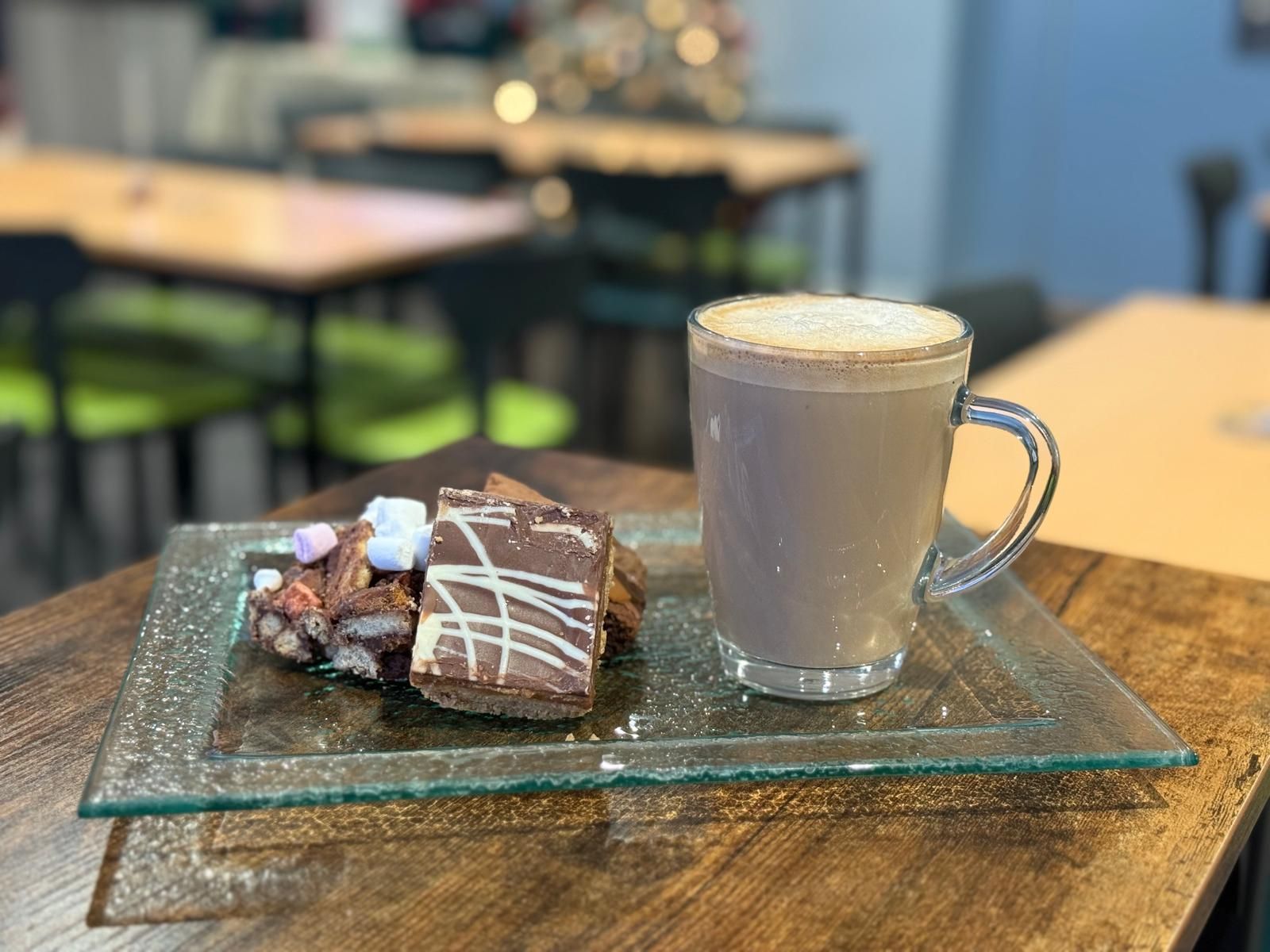 Hot chocolate with froth, and rocky road treats on a glass plate, on a wooden table, in a cafe.