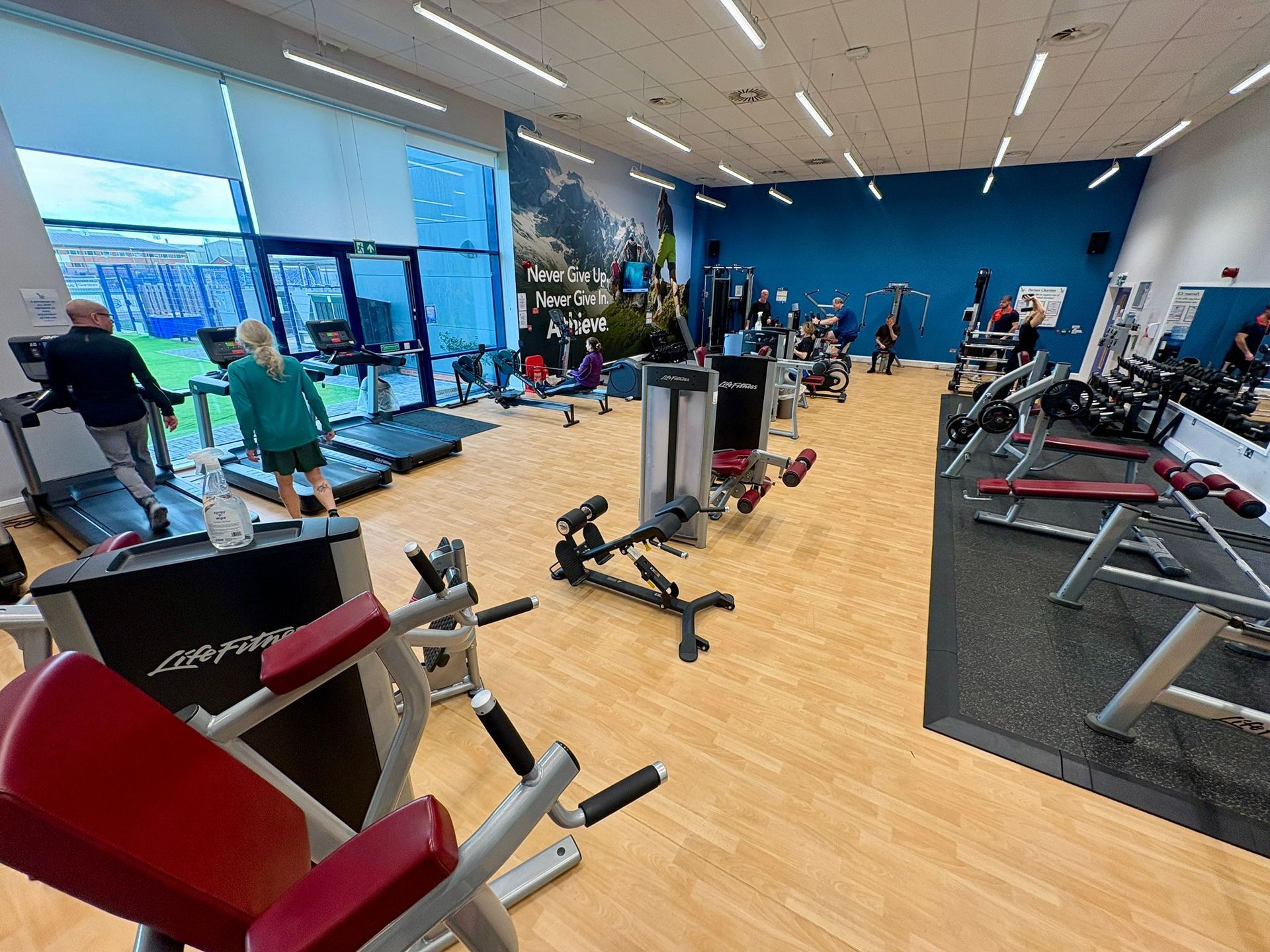 Gym interior with fitness machines and people exercising. Sunlight streams in from a large window.
