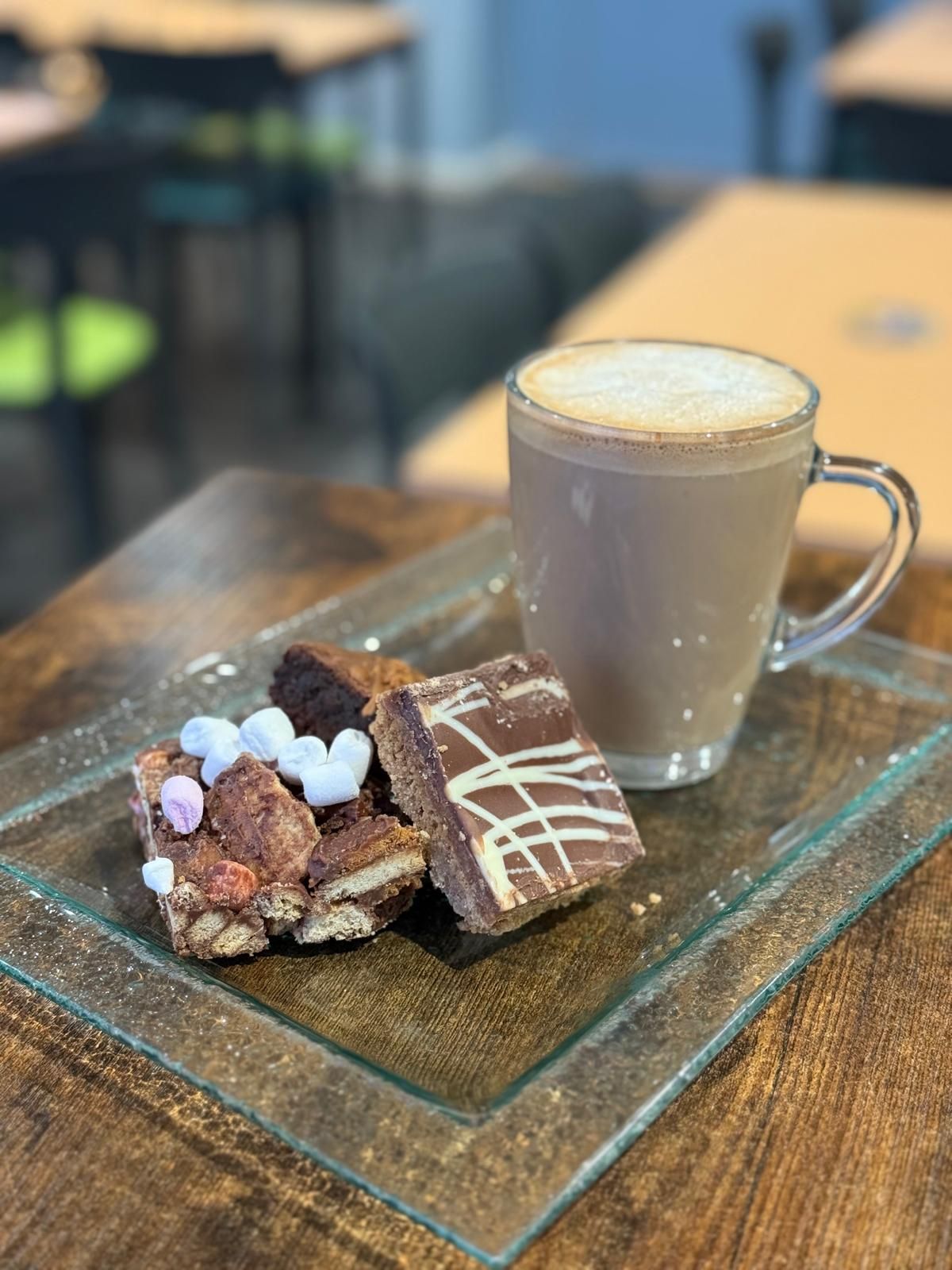 Hot chocolate with foam, and various chocolate treats on a glass plate on a wooden table.