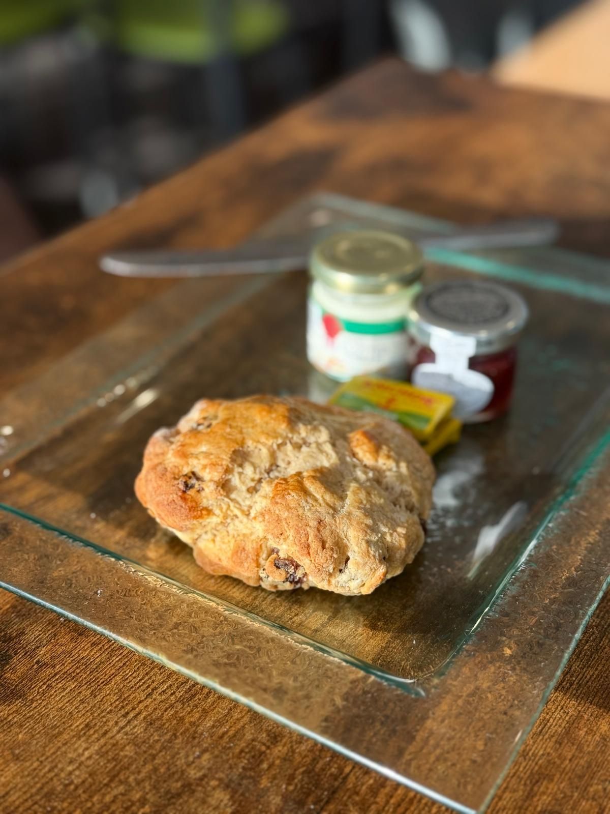 Scone with jam, butter, and cream on a glass plate. Wooden table in background.