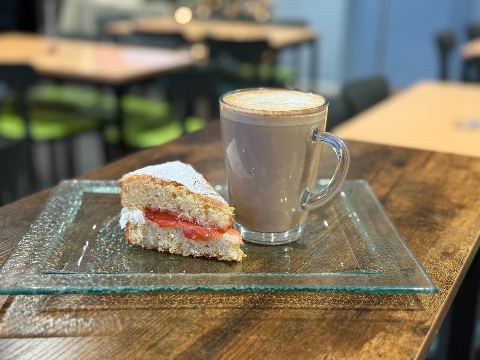 Slice of cake with a strawberry filling and a latte in a glass mug on a glass plate. Wooden table.