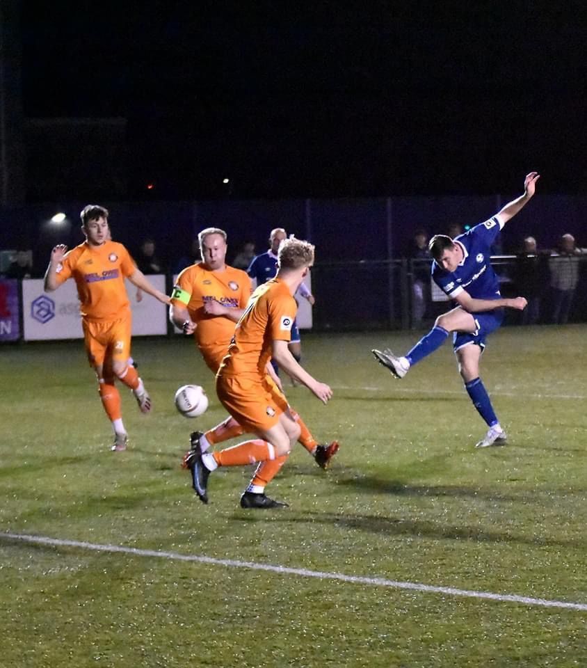 Soccer player in blue kicks the ball, orange-clad players attempt to block, night game.