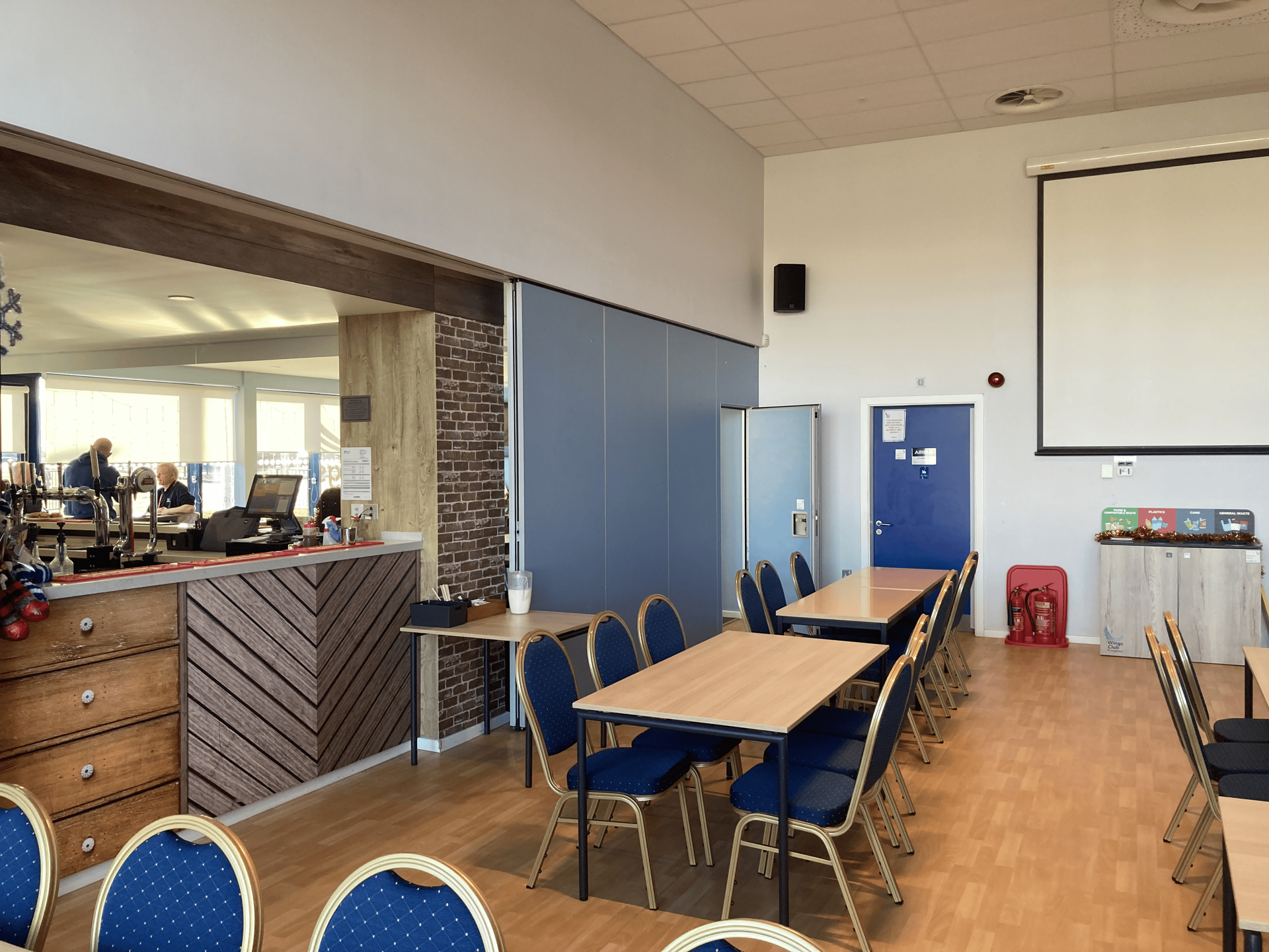 Interior of a meeting room with tables, chairs, a projector screen, and a serving area with people visible.