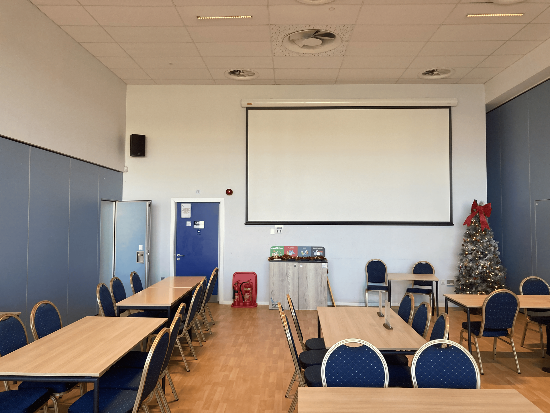 Empty classroom with long tables, blue chairs, projector screen, and a small Christmas tree.