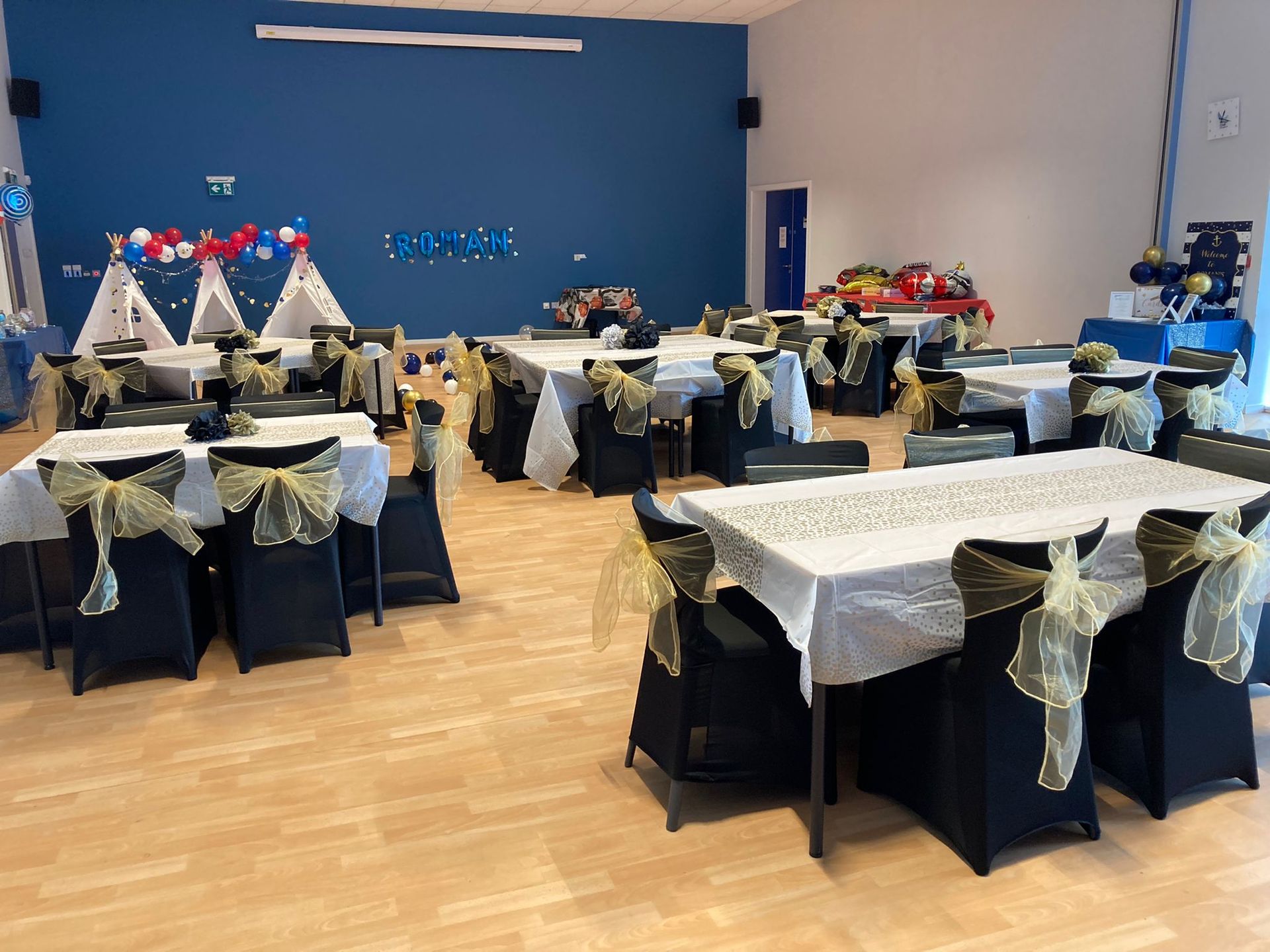 Event hall set for a party, tables draped in white, black chairs with gold bows, blue walls, decorations.