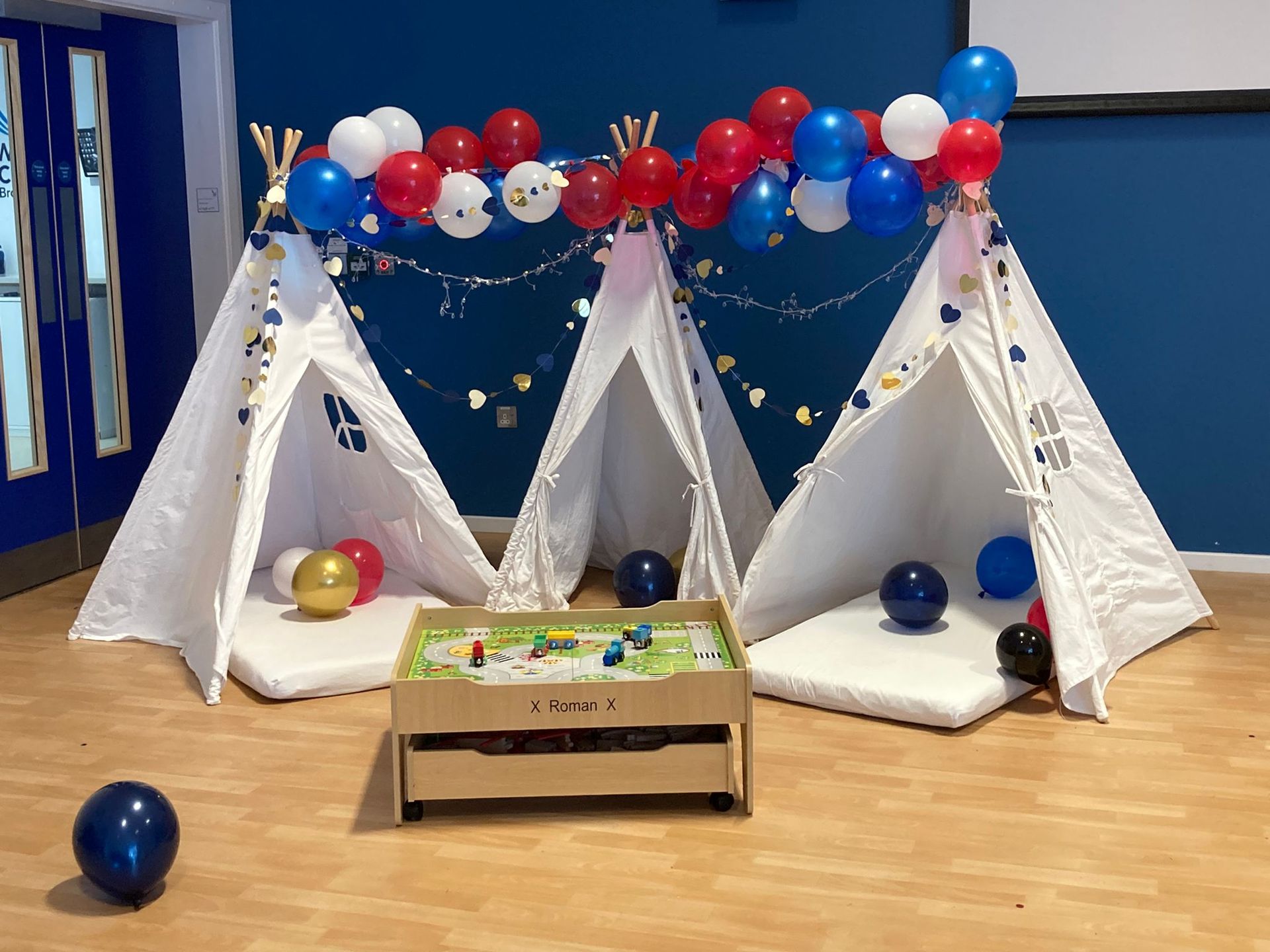 Three white teepees decorated with balloons, lights, and gold stars, with a wooden table in front.