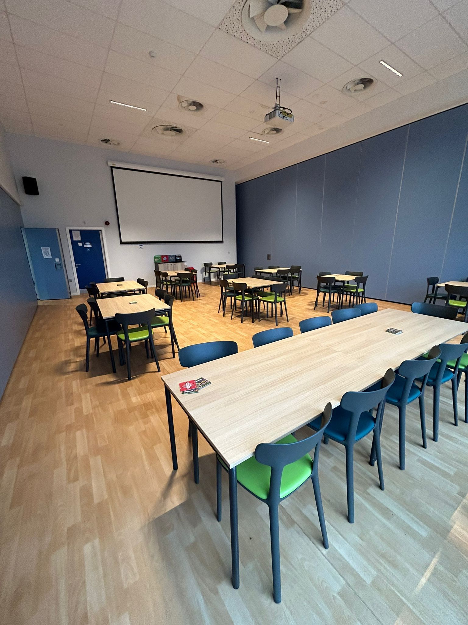 A classroom with square tables, chairs, and a long table. Blue wall with projector screen.