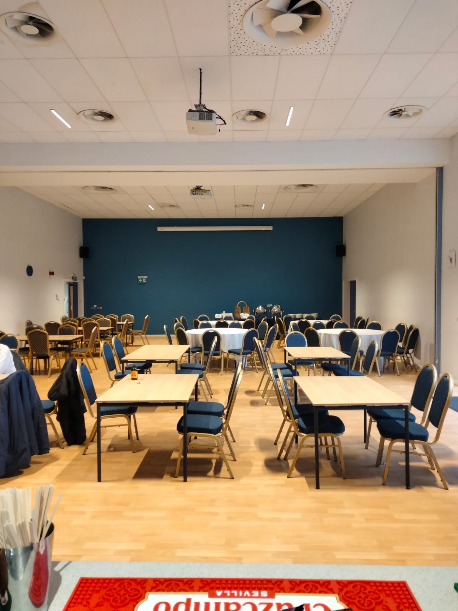 Empty event hall with tables, chairs, and a blue accent wall. Lighting and projector on the ceiling.