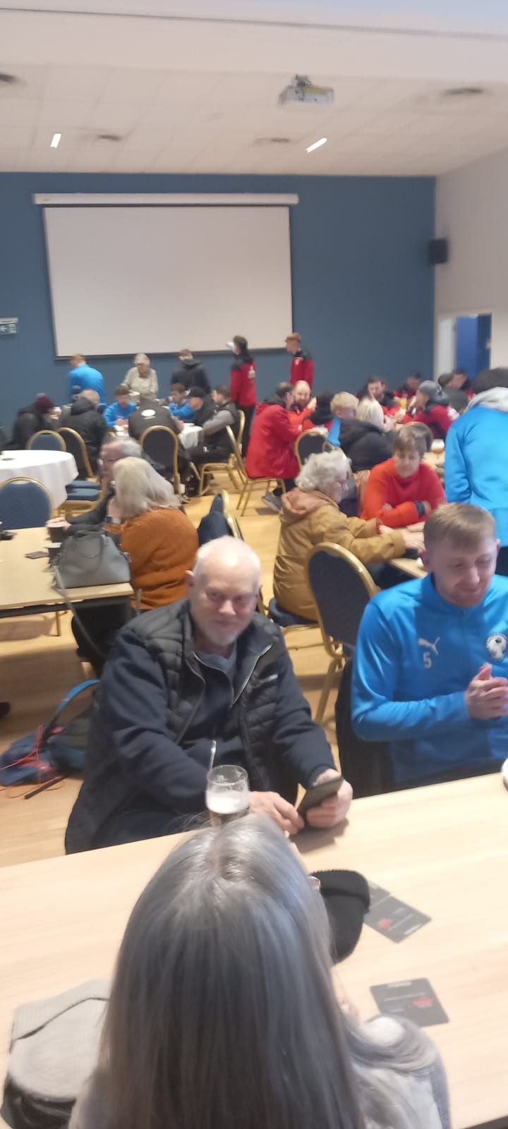 A crowded room with people seated at tables, some are looking at a presentation on a screen.